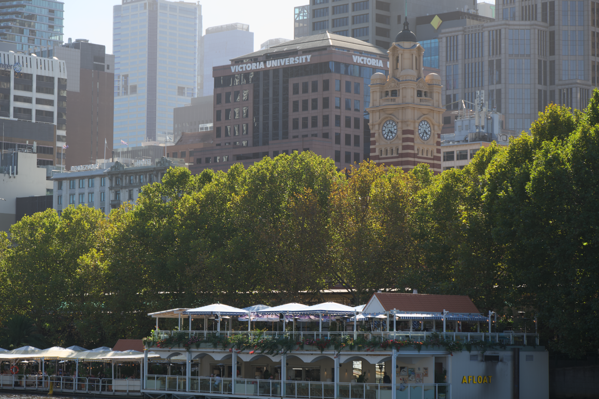 A cityscape with a mix of modern skyscrapers and a historic clock tower, with green trees and an outdoor dining area with umbrellas in the foreground.