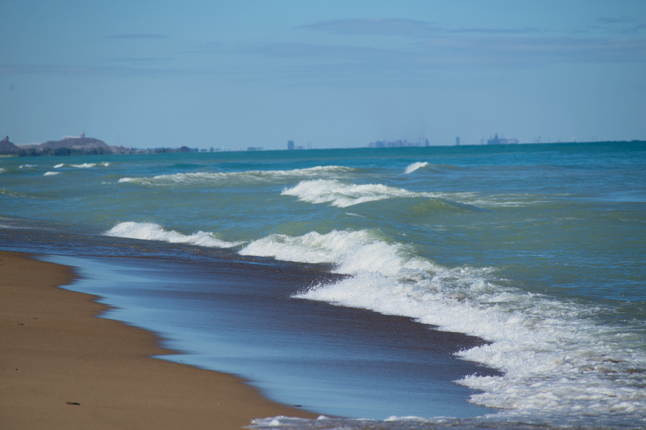 Waves crashing on a sandy beach with a city skyline in the distance under a partly cloudy sky.