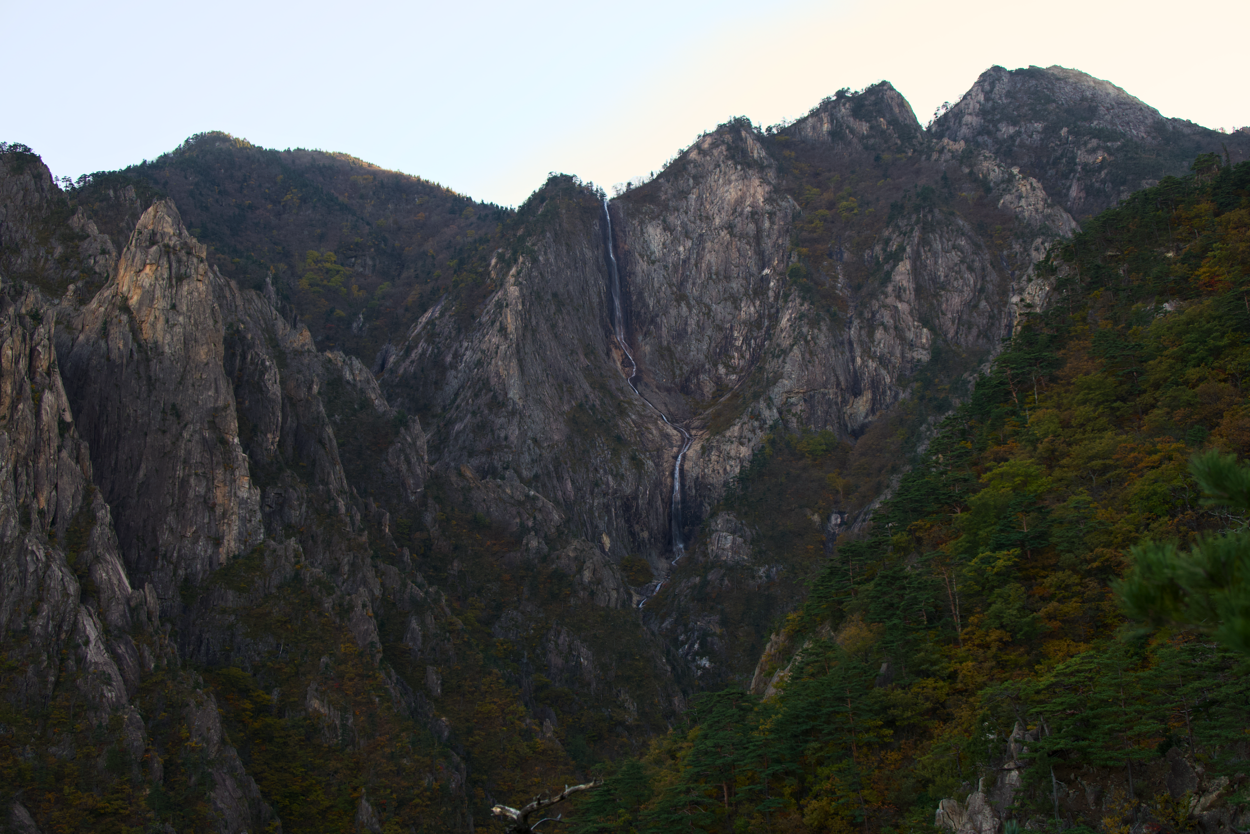 Mountains with rocky cliffs and a waterfall, surrounded by trees with autumn foliage, under a clear sky.
