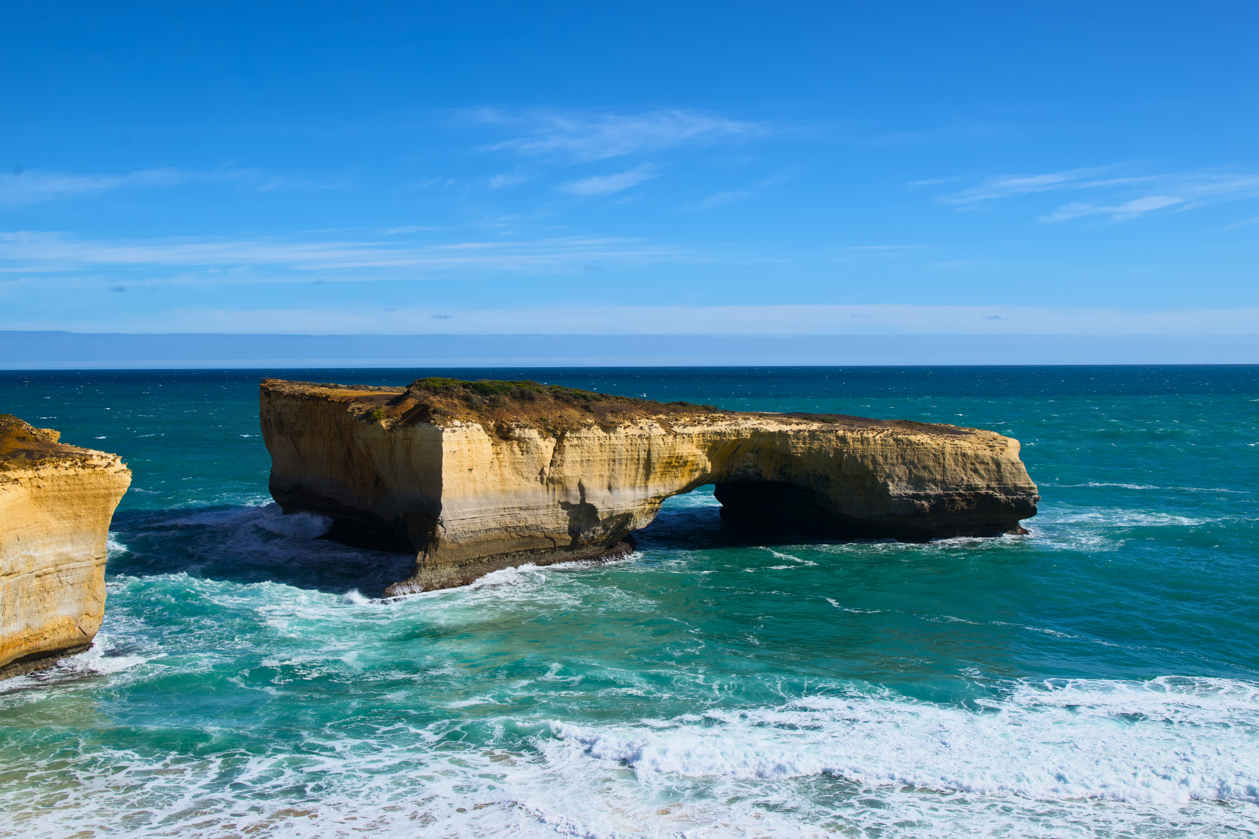 Bright blue sky over a sea stack with a natural arch in the ocean, surrounded by turquoise water and white waves.