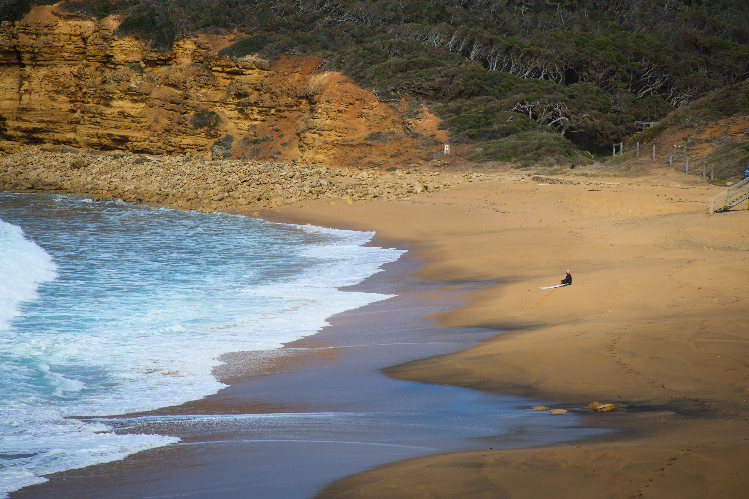 A person sitting on the sand at a beach with cliffs and greenery in the background, gentle waves washing onto the shore.