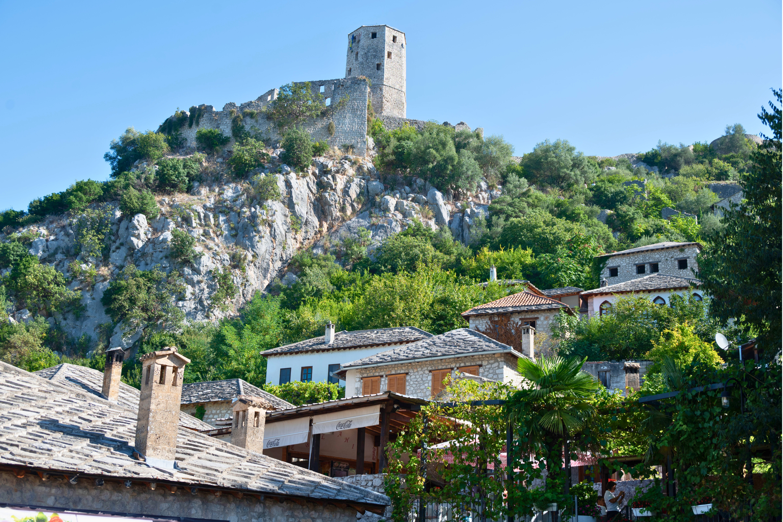 A hillside village with stone houses and a castle on top of the hill, surrounded by lush green trees and clear blue sky.