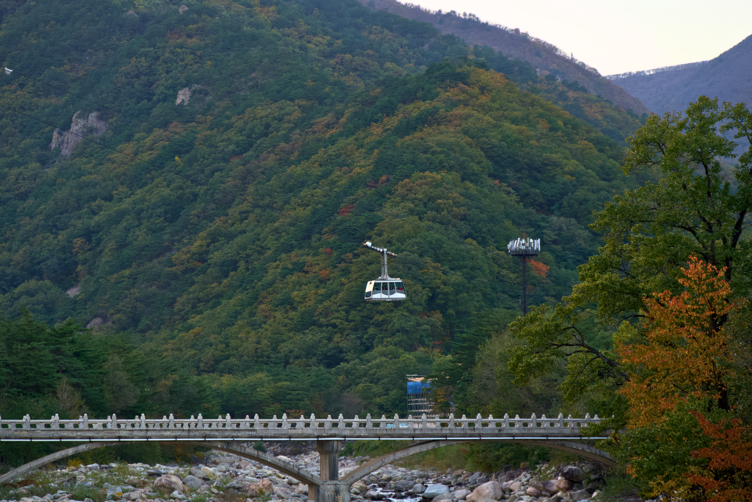 A cable car floating in front of a forested mountain with green, orange, and yellow foliage, and a stone bridge over a rocky river in the foreground.