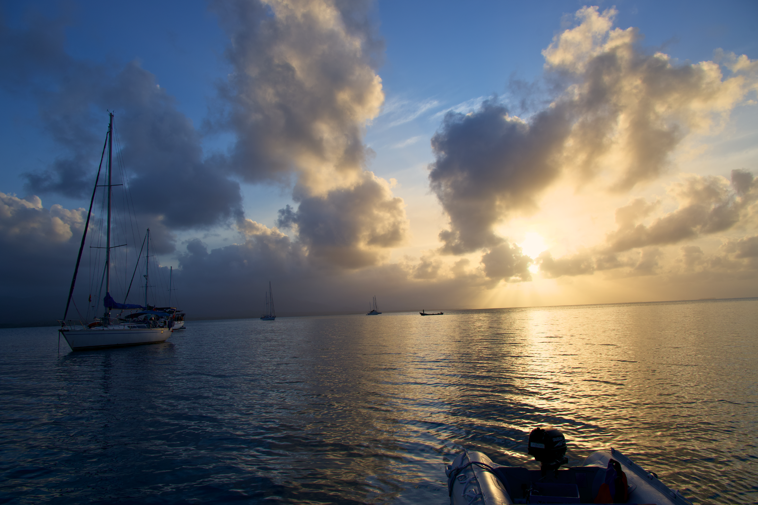 A scenic view of a sunset over calm water with boats anchored, including part of an inflatable boat in the foreground, and partly cloudy sky.