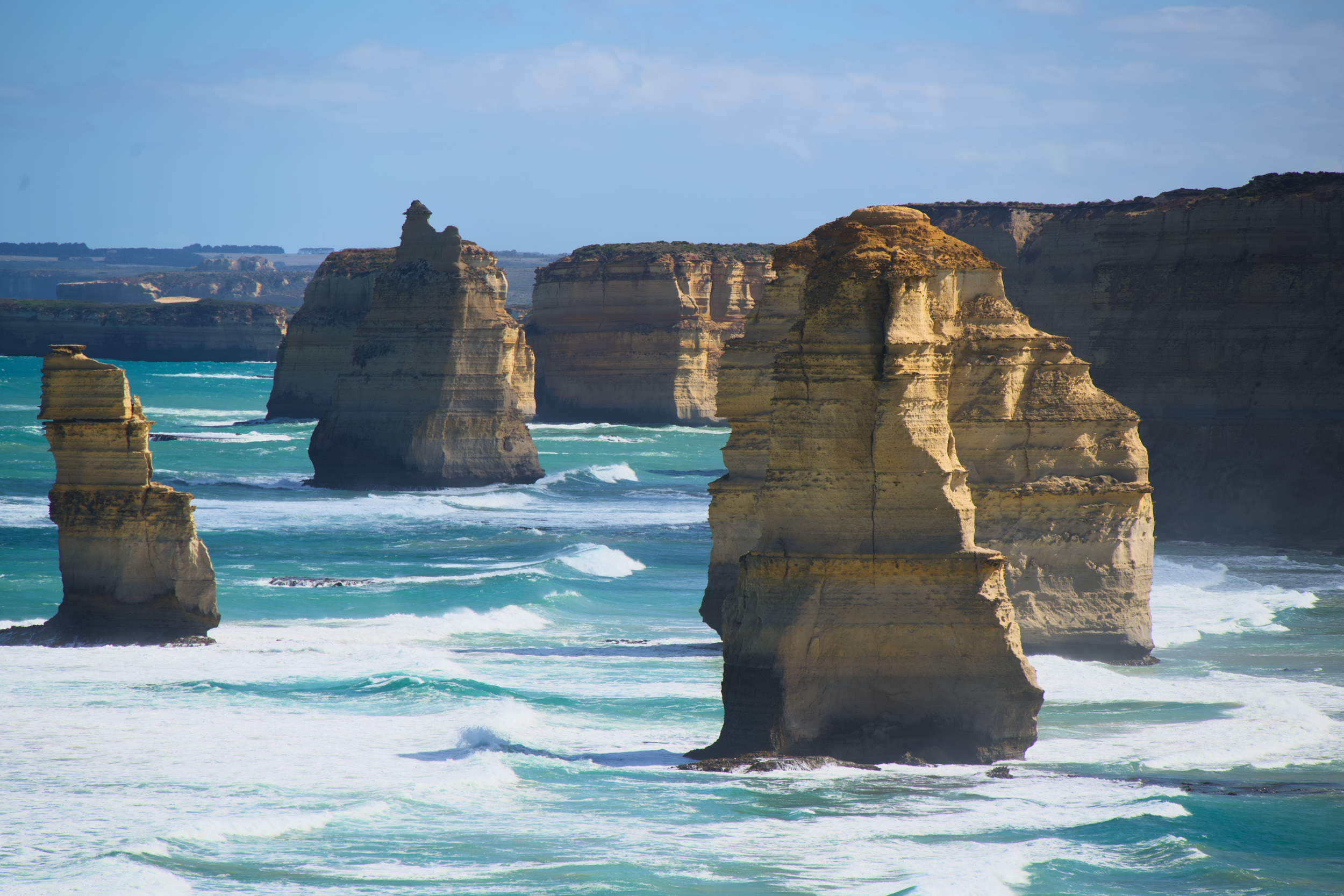Cliff formations along the coastline with ocean waves crashing at the base, under a partly cloudy sky.