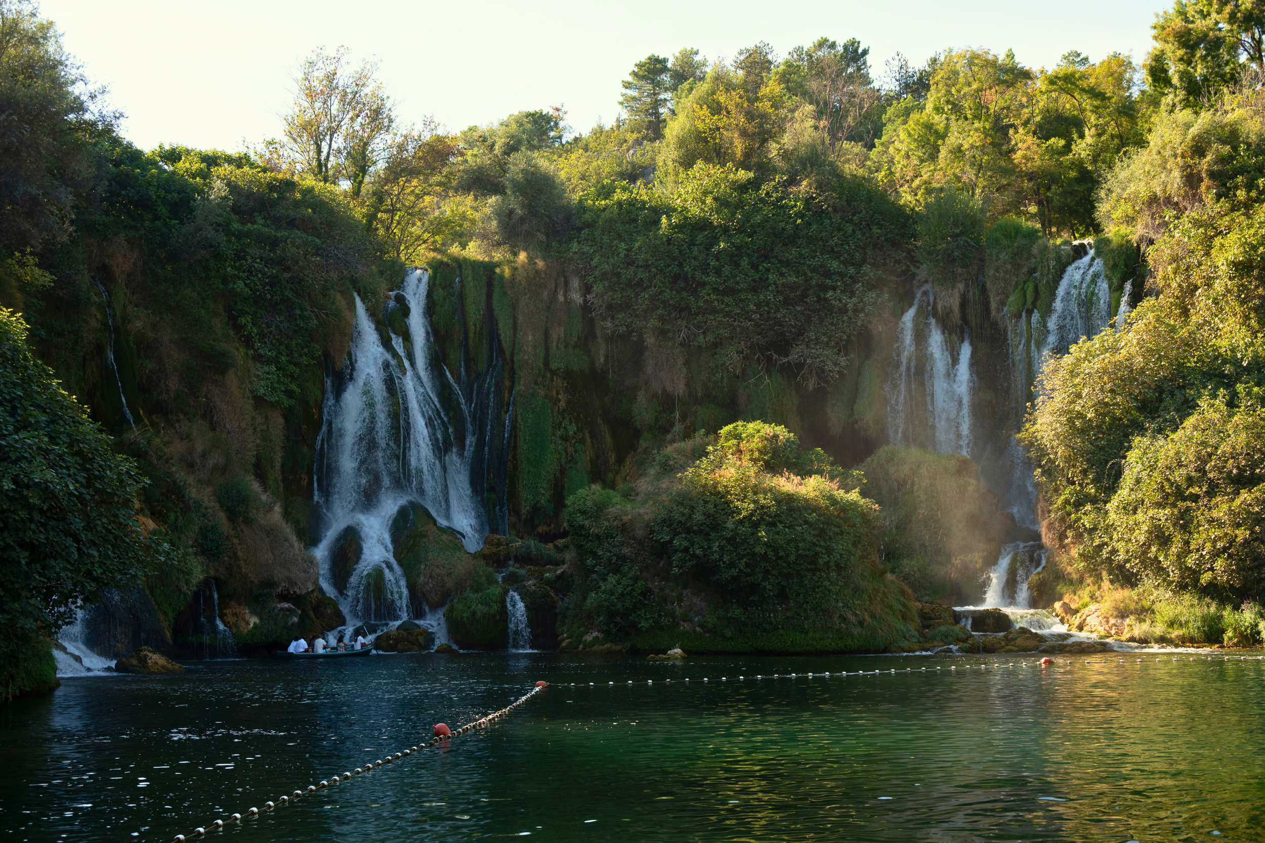 Waterfalls flowing into a large pond, surrounded by lush green trees, with a group of people in a boat near the base of the falls.