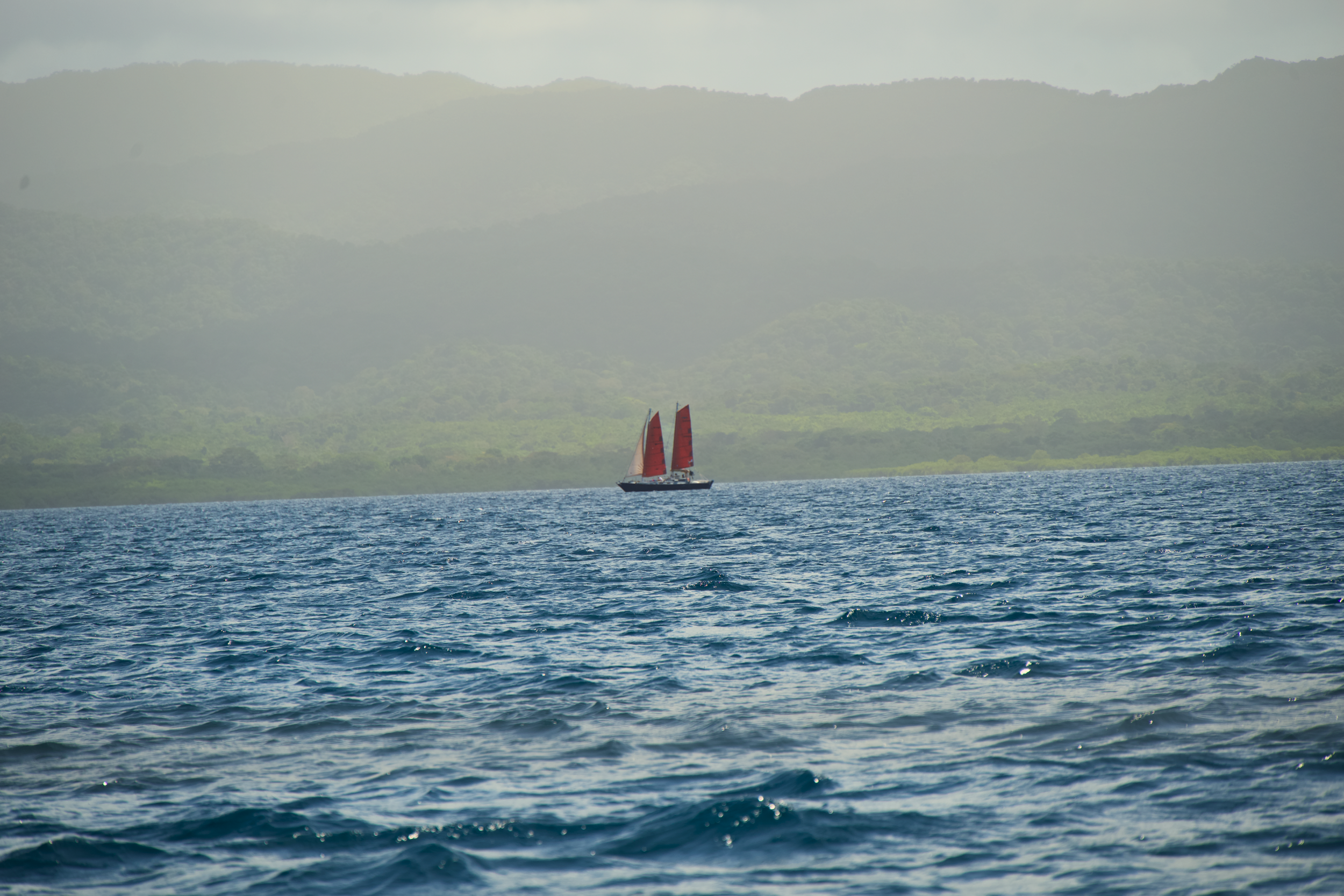 A sailboat with red sails on the ocean, with green mountains in the background under a hazy sky.