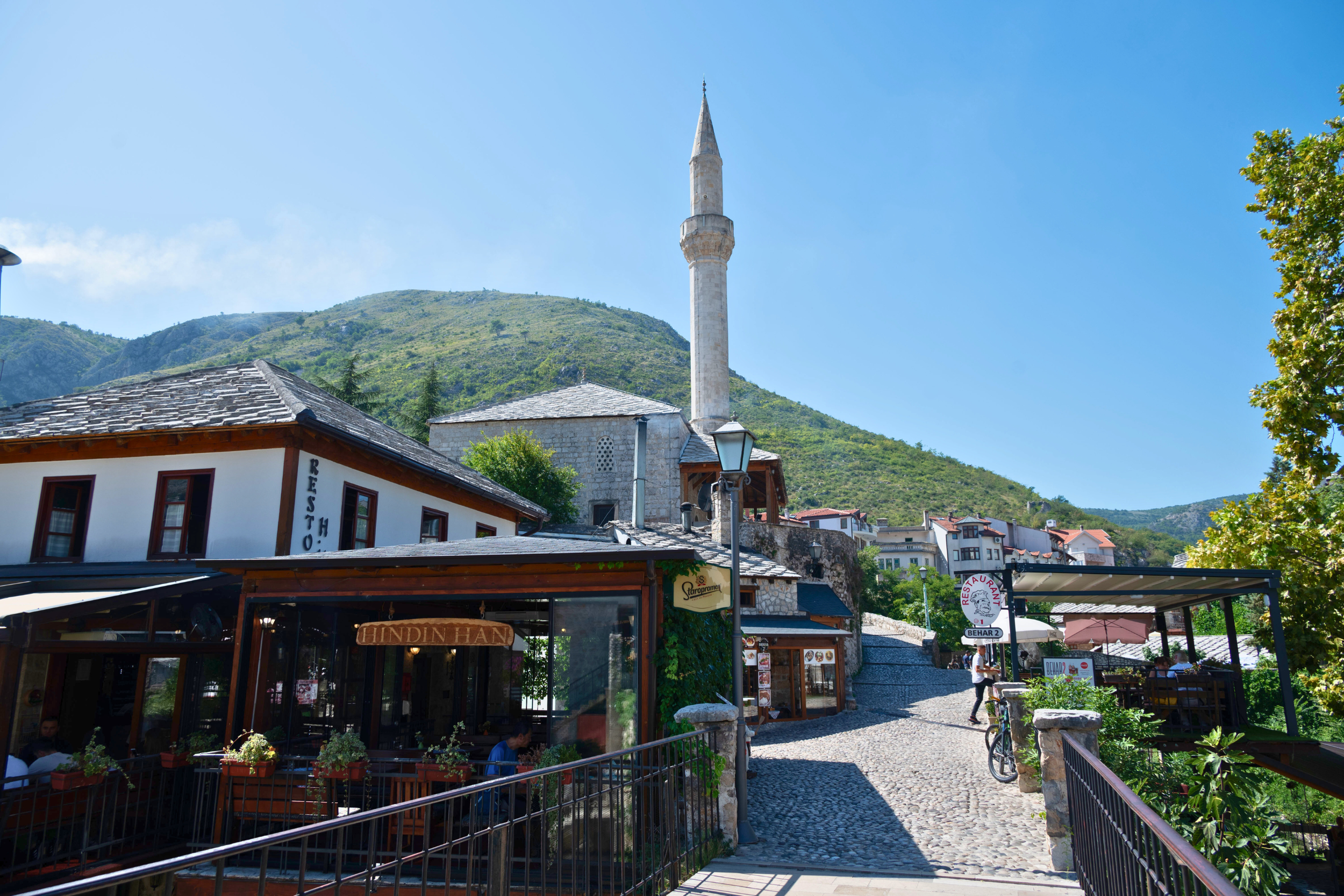 A scenic view of a small town with a mosque featuring a tall minaret, surrounded by green mountains and blue sky. In the foreground, there are cafes and shops with outdoor seating and signs, cobblestone paths, and people walking or sitting.