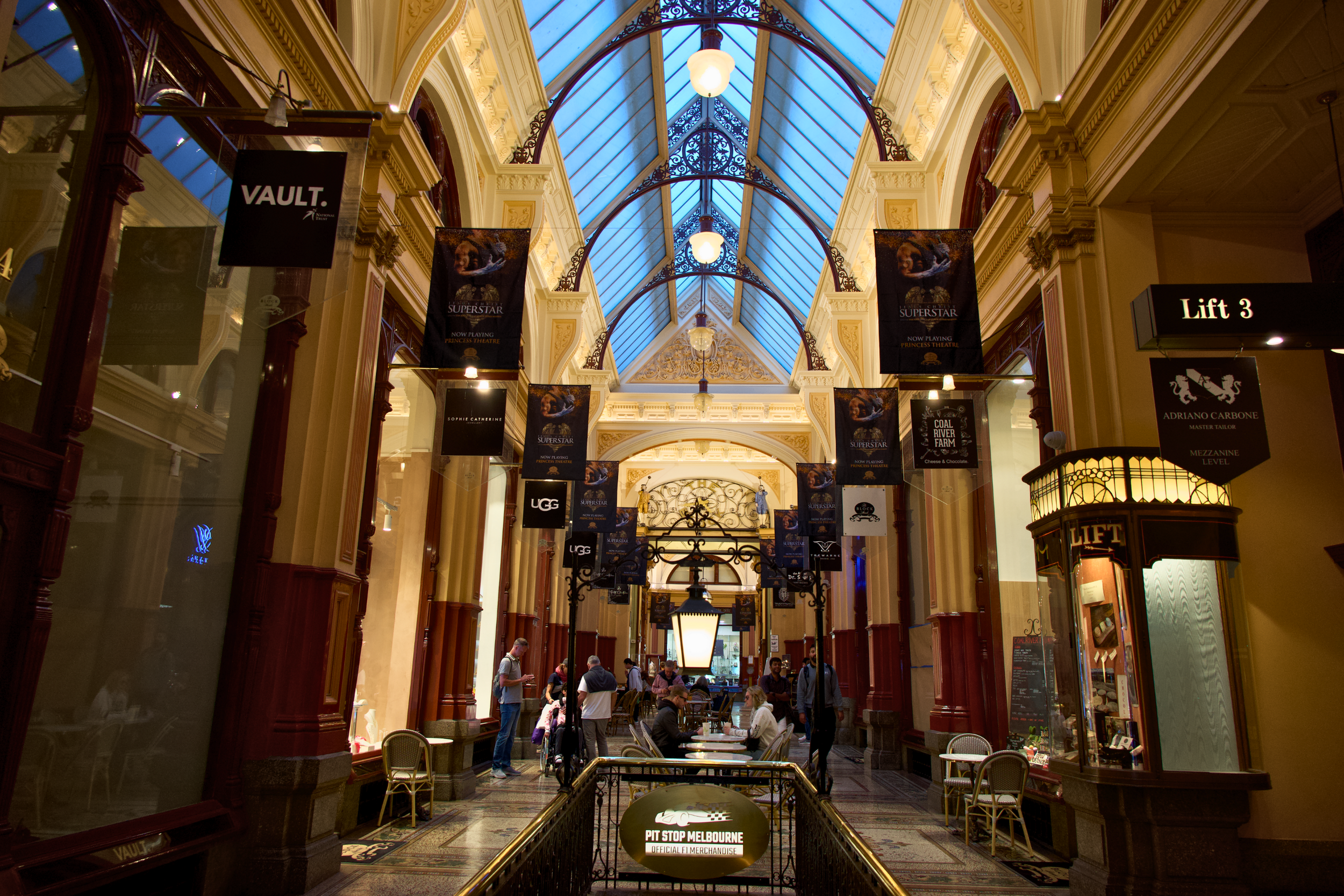 Interior of a shopping arcade with a glass ceiling, hanging signs, and people sitting and walking.