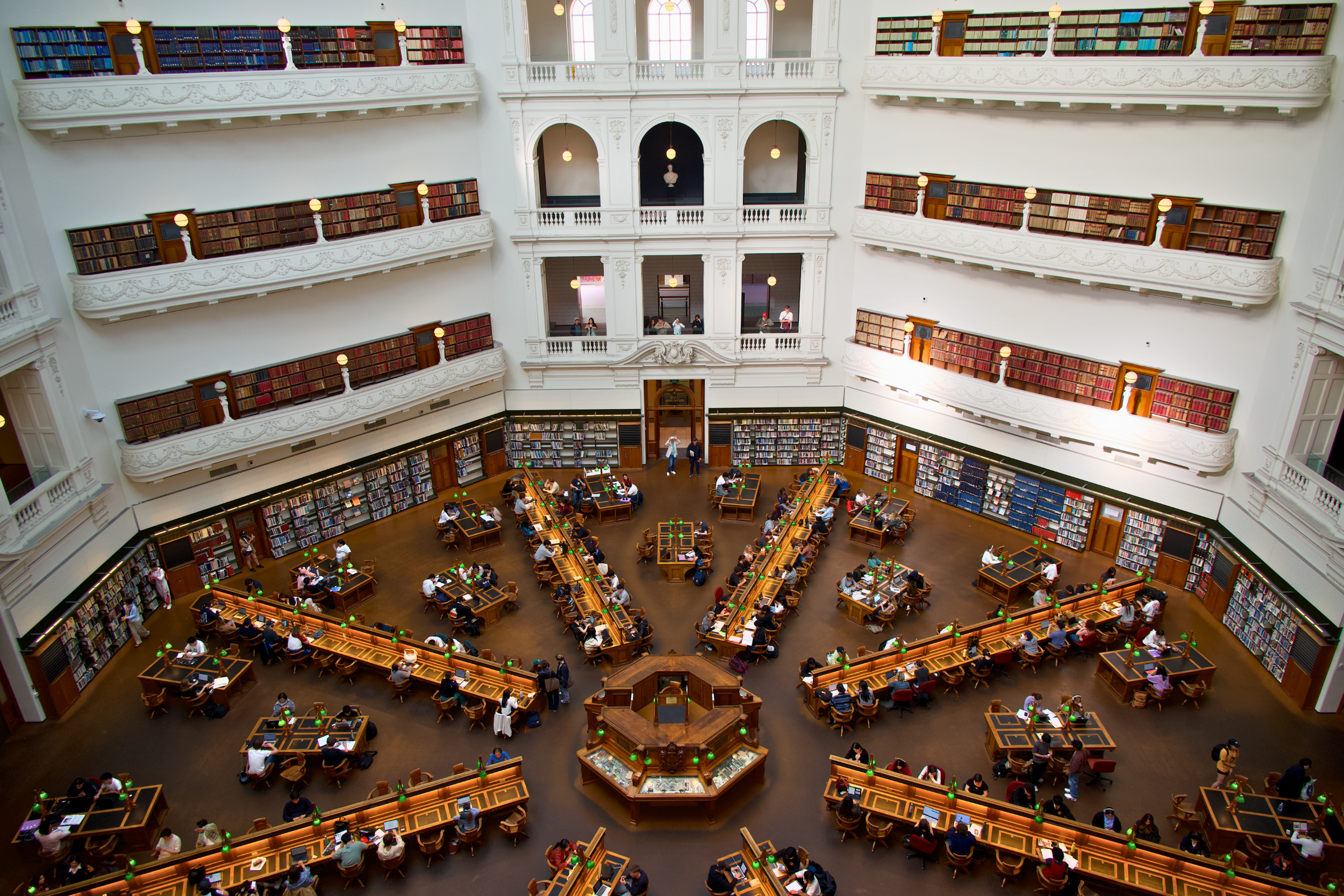 A large, ornate library with white walls, multiple levels of bookshelves, and a high ceiling. The main floor features long wooden tables with green reading lamps and numerous visitors reading or working. The upper balconies also have bookshelves and some visitors looking over the railing.