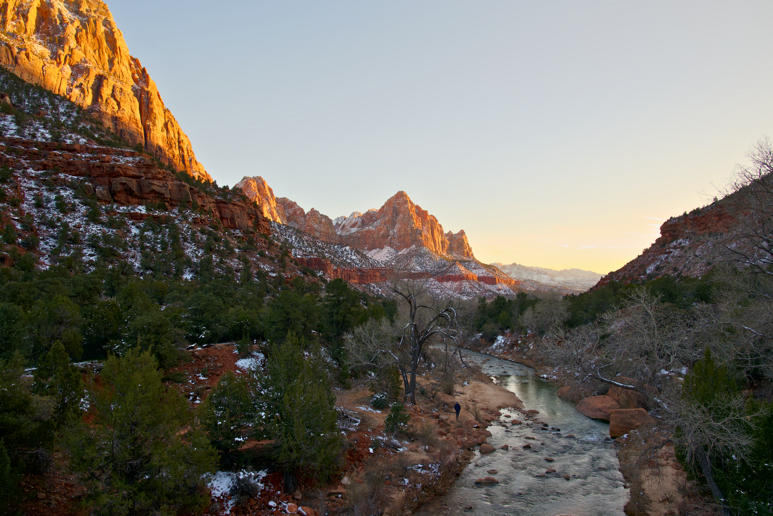 Scenic view of Zion National Park with rugged red cliffs, sparse snow patches, green trees, and a stream at sunset.