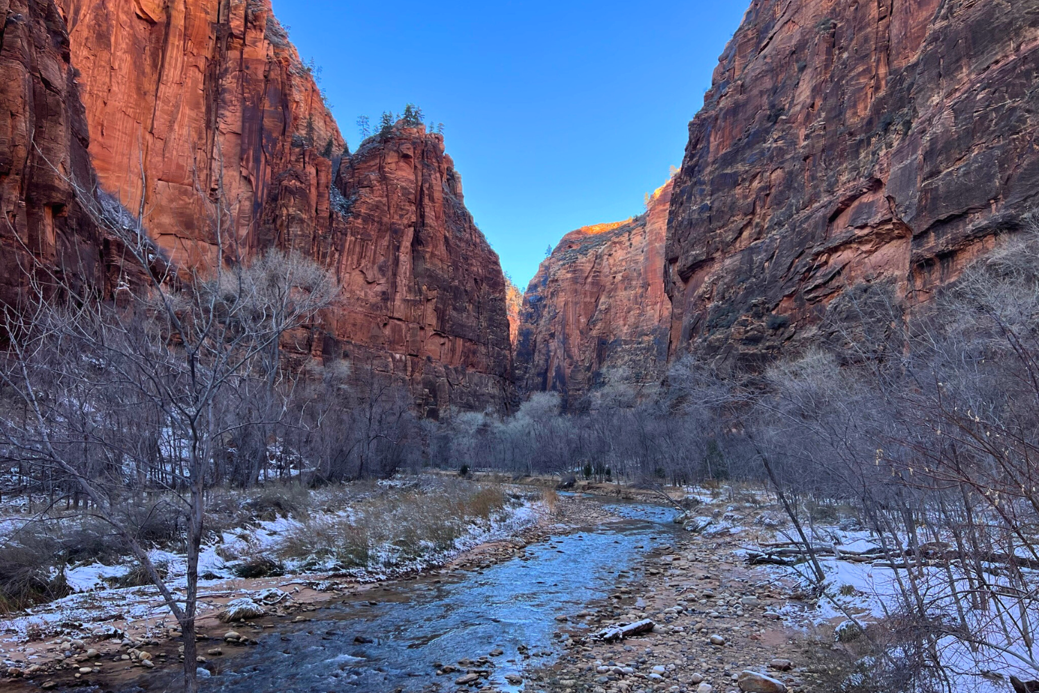 View of a narrow canyon with tall red rock cliffs on either side, a small river flowing through the rocky landscape, and leafless trees along the banks, with patches of snow on the ground and a clear blue sky overhead.
