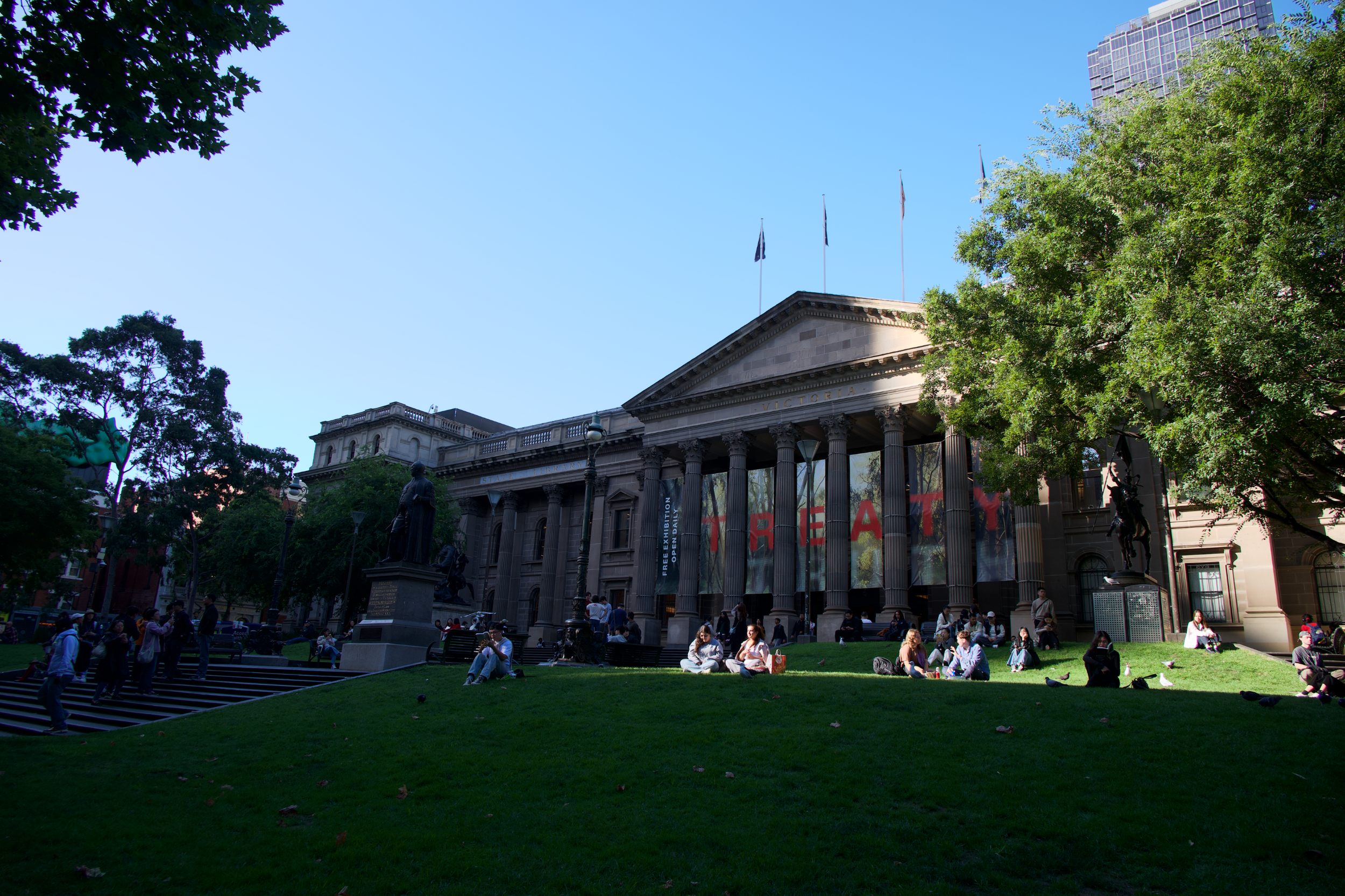 People sitting and walking on a grassy area in front of a historic courthouse with large columns, statues, and a banner with the word 'CREATY' on it, surrounded by trees and modern buildings.
