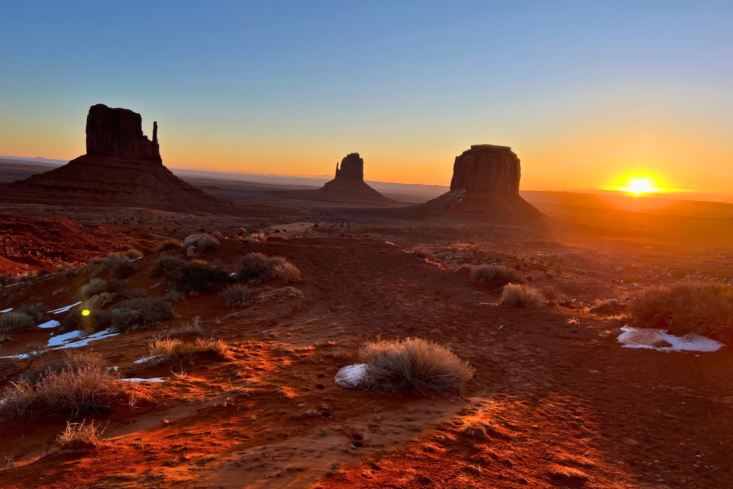 Sunset over Monument Valley with three large sandstone buttes rising from a desert landscape, sparse vegetation, and a clear sky.