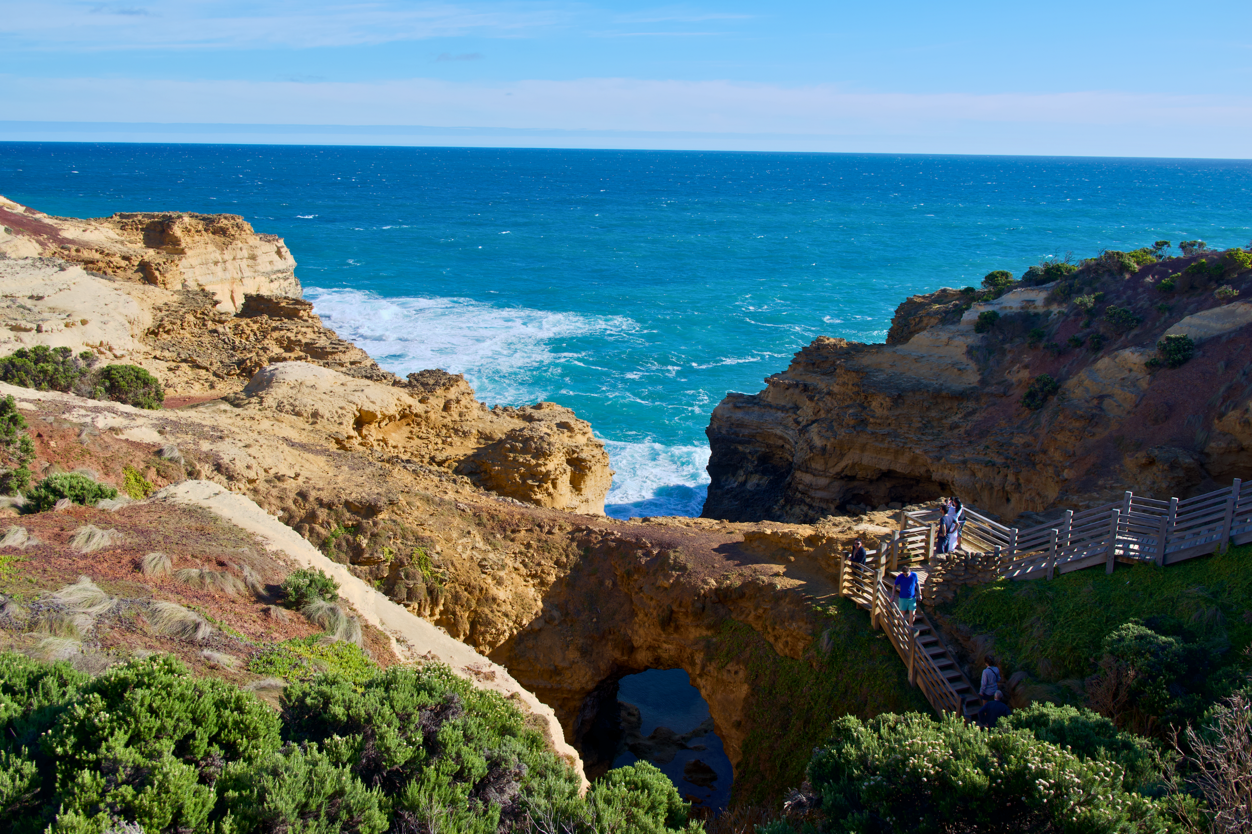 Cliffside coastline with a natural arch, ocean waves, and a wooden staircase with people walking on it