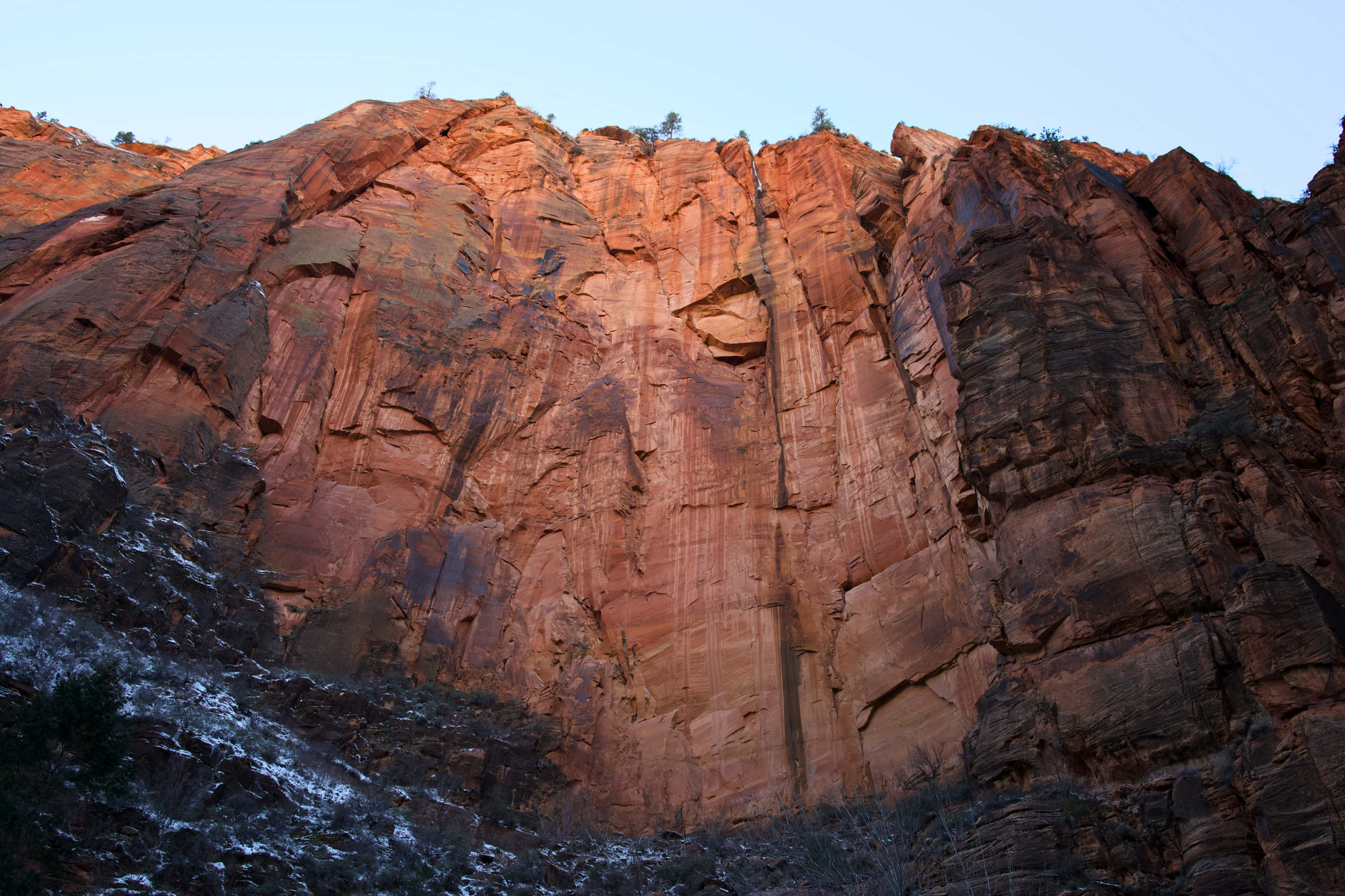 Tall, reddish-orange rock cliffs with layered striations in a desert landscape. Sparse vegetation and snow on the ground at the base, with a clear light blue sky above.