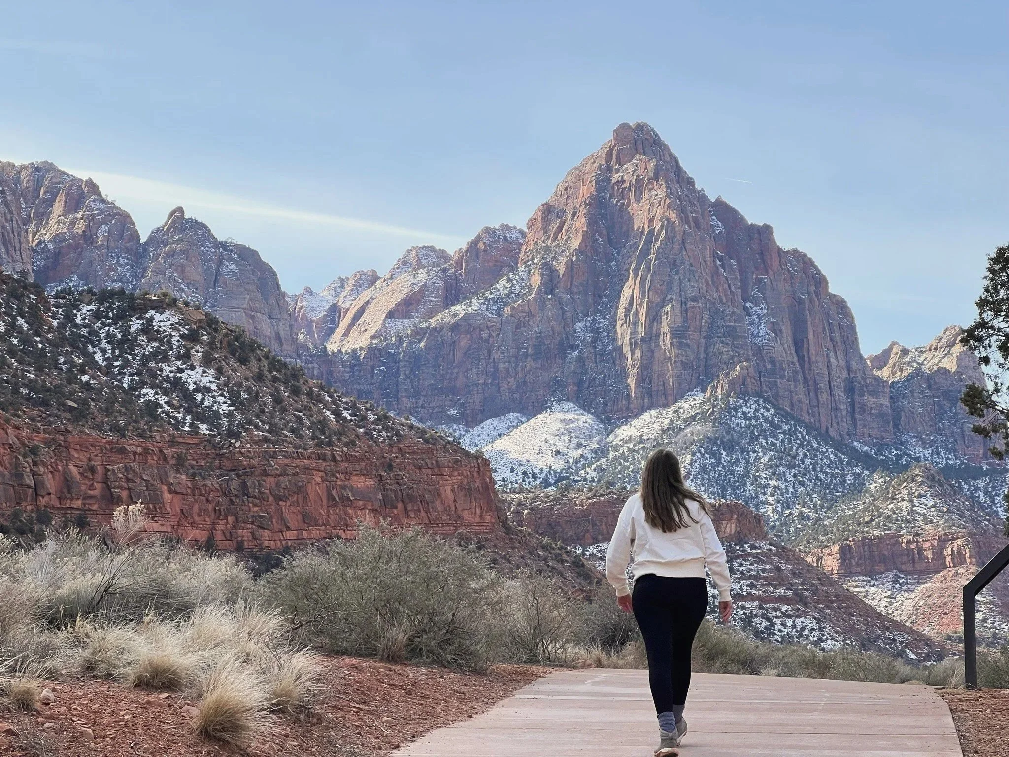 A woman walking on a paved trail in a mountainous area with snow-capped peaks and red rock formations in the background.