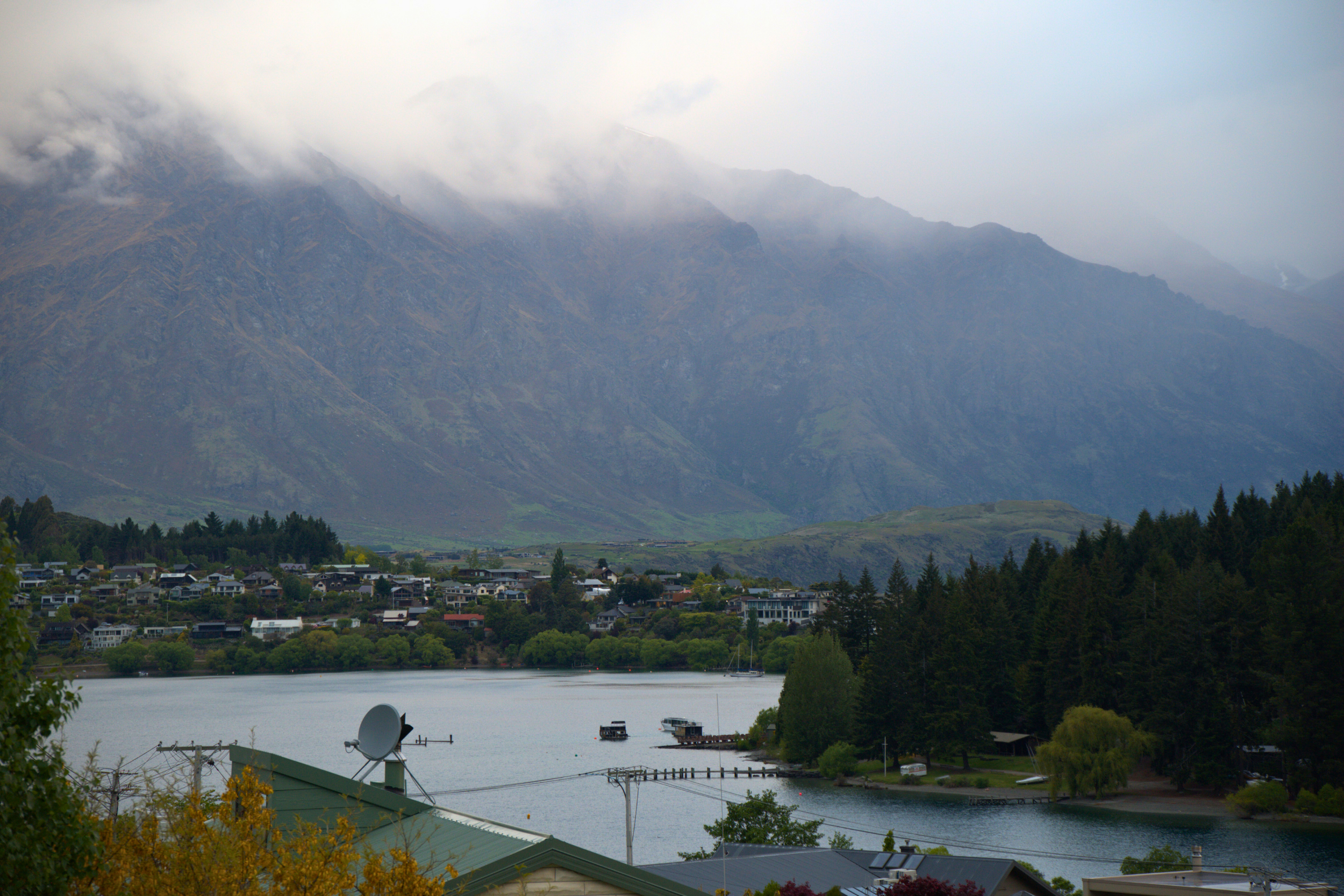 A lakeside town with houses near the water, surrounded by green trees and dense forest. In the background are tall mountain ranges partially covered by clouds.
