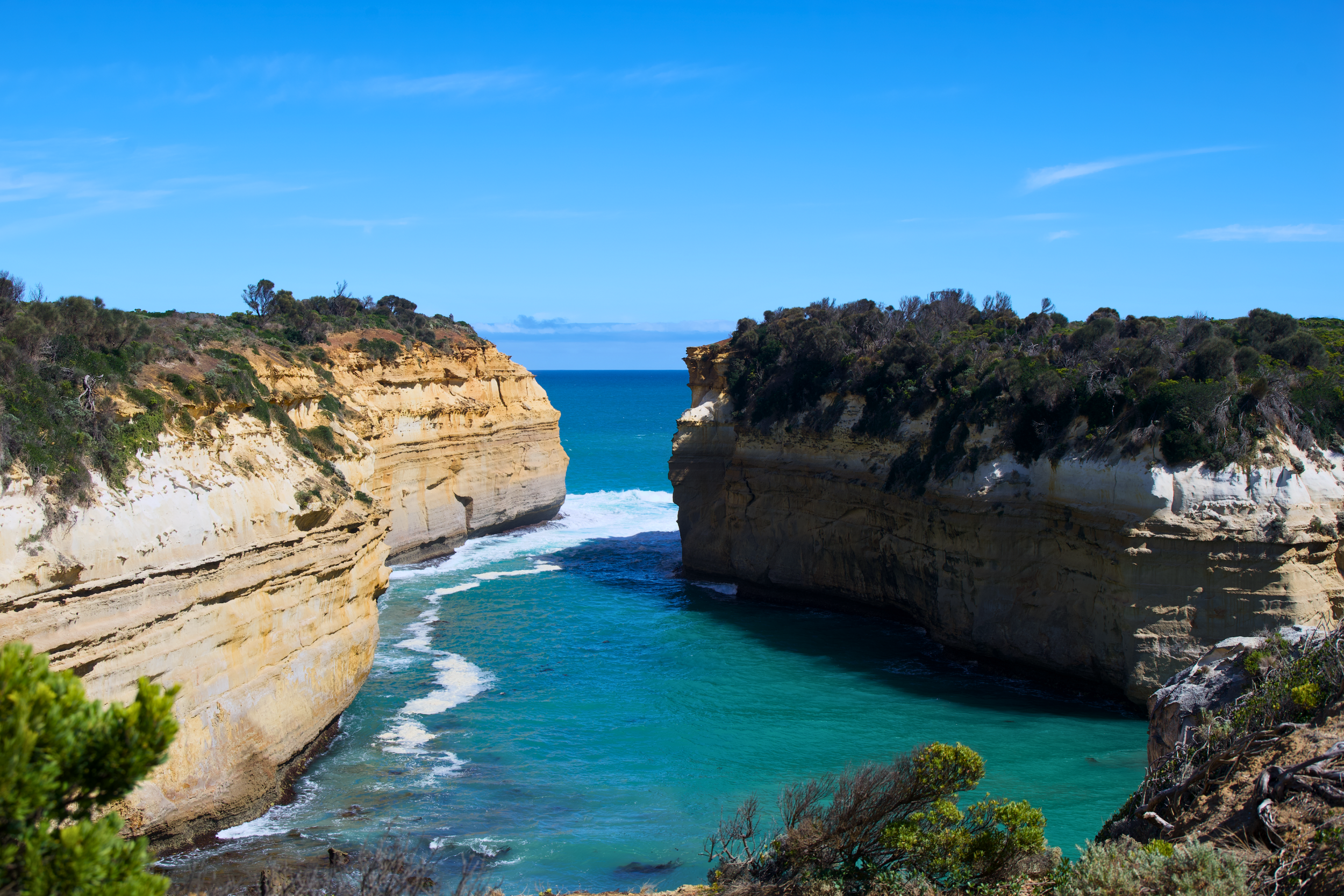 Photo of a coastal landscape with large sandy cliffs on either side of a narrow blue water channel, under a clear blue sky.