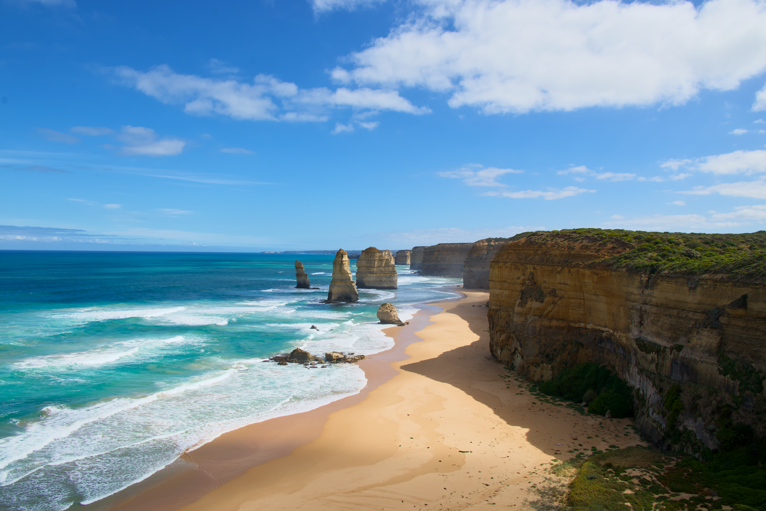 Beach with tall rock formations in the ocean along a sandy shoreline, cliffs covered with green vegetation, blue sky with scattered clouds.