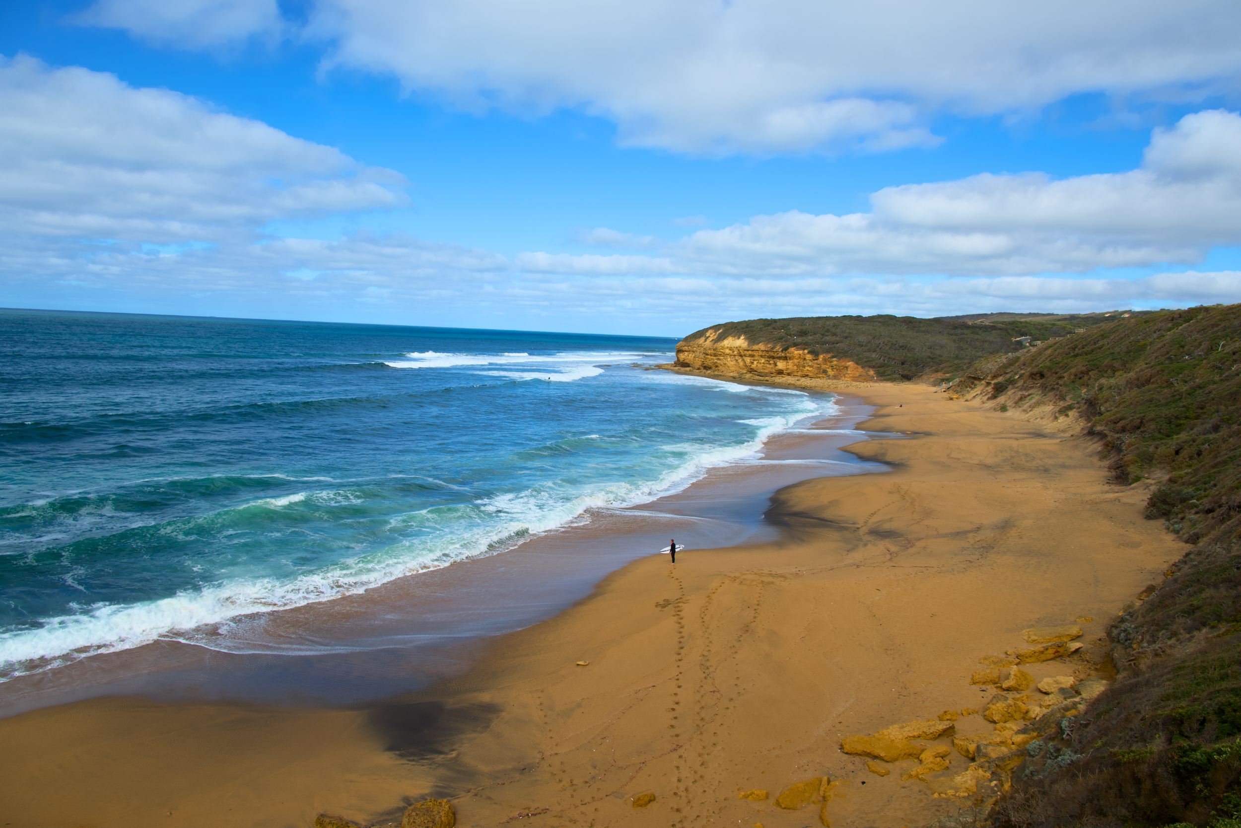 A person walking along a sandy beach with waves crashing, surrounded by cliffs and a partly cloudy sky.