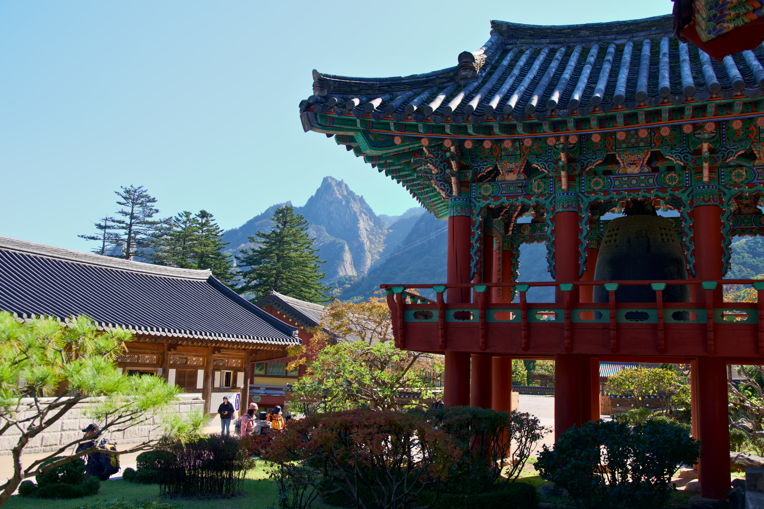 Traditional Asian temple with a colorful, ornate roof, set against a mountainous landscape with trees and clear blue sky.
