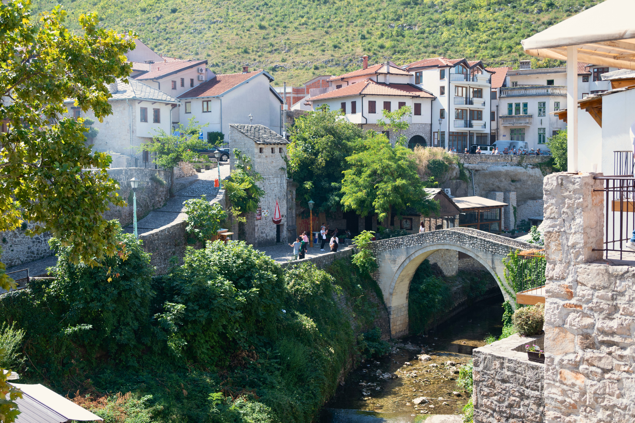 A scenic view of a small town with white buildings, a stone bridge over a river, and green trees, under a hillside on a sunny day.