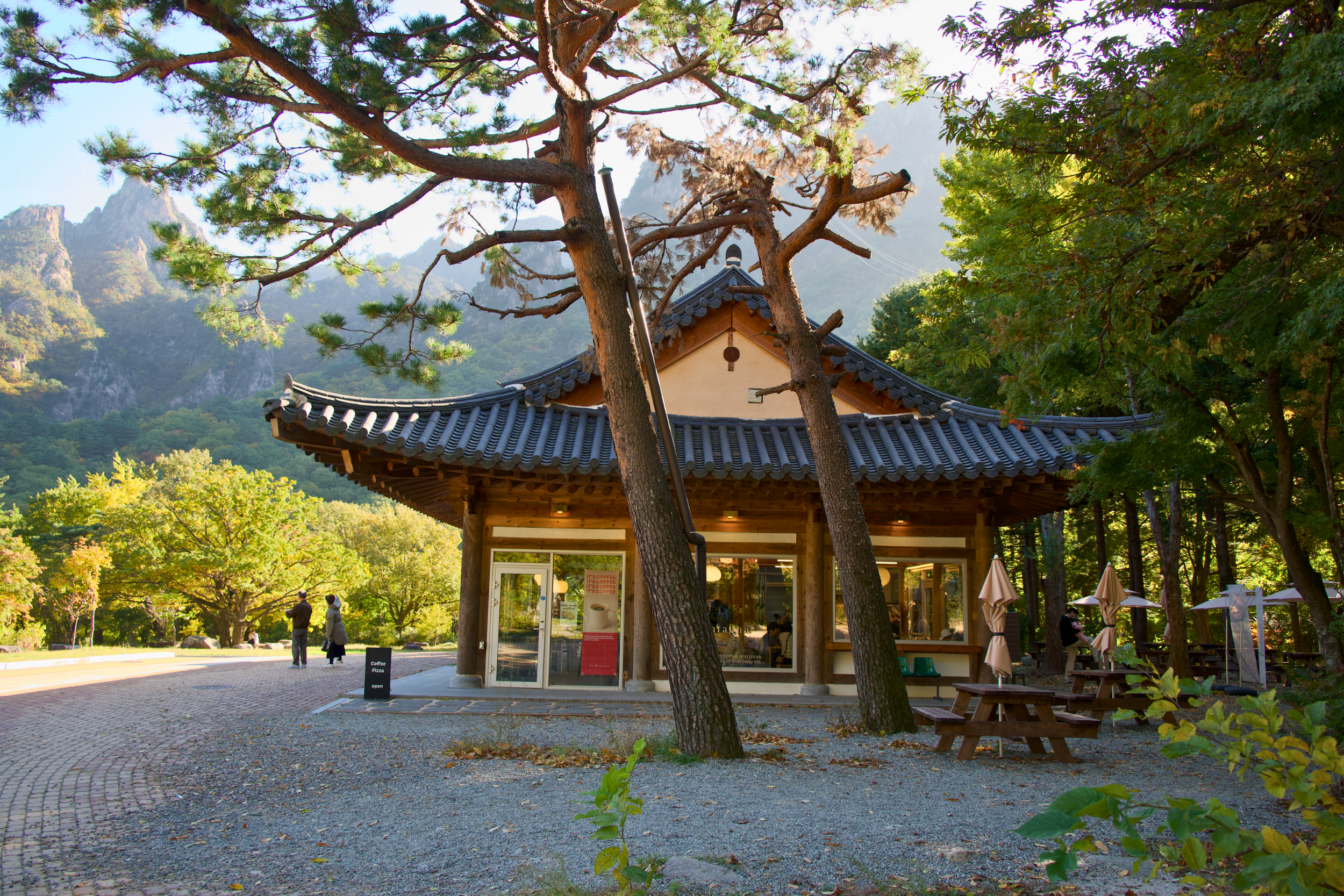 A traditional Asian-style building with a tiled roof, surrounded by trees and green foliage. There are people standing and sitting outside, and the background features mountains and a clear sky.