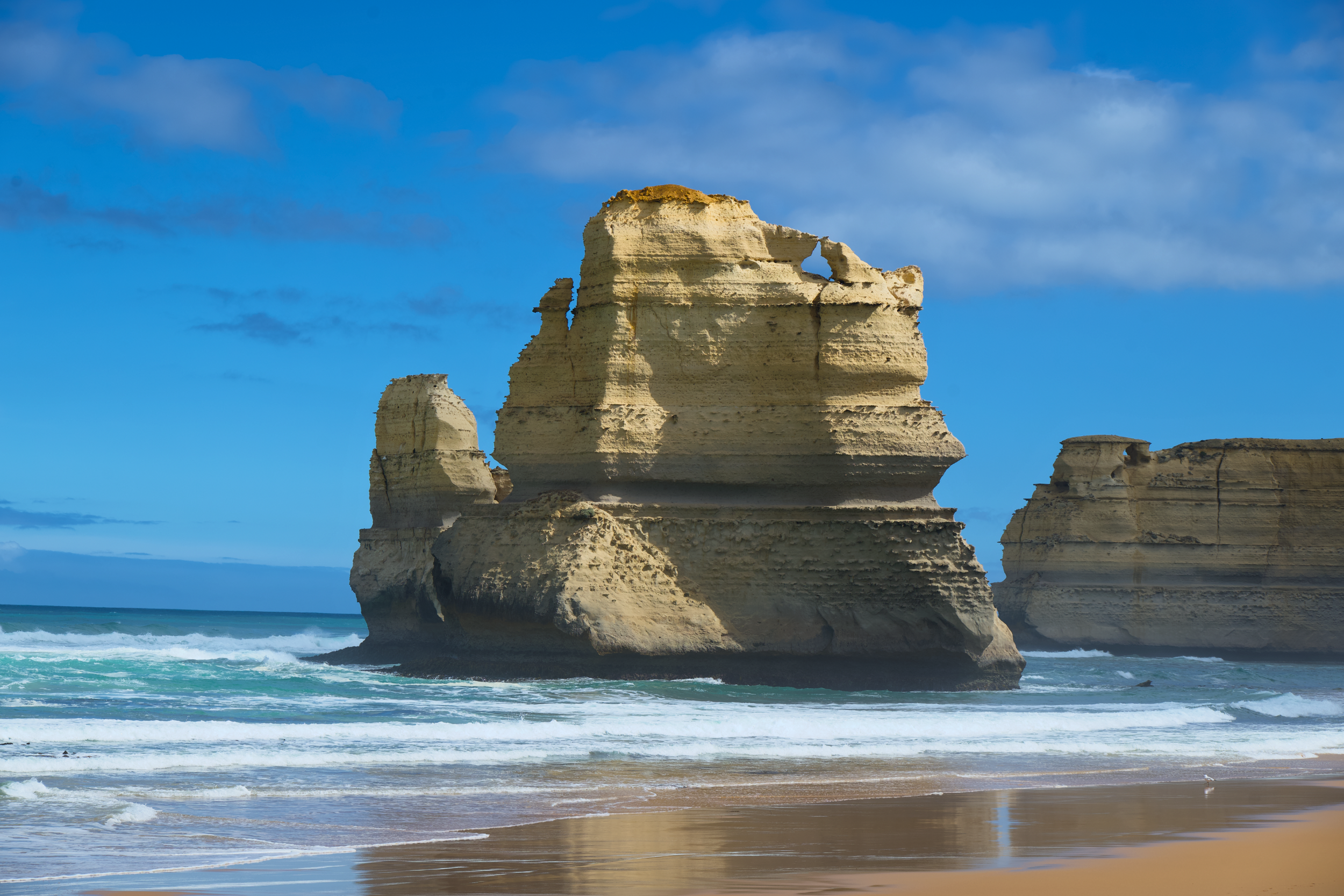Large beige rock formations along a sandy beach with ocean waves and a partly cloudy blue sky.