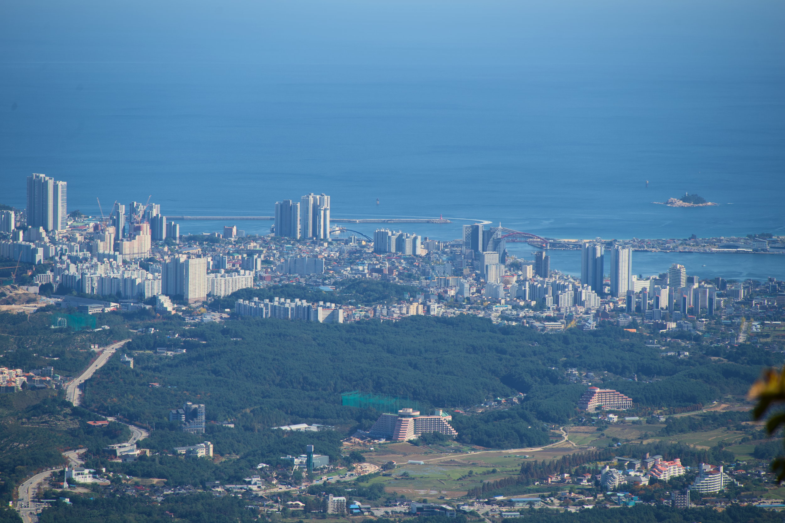A cityscape with tall buildings, a bridge over water, and an island with a lighthouse in the ocean.
