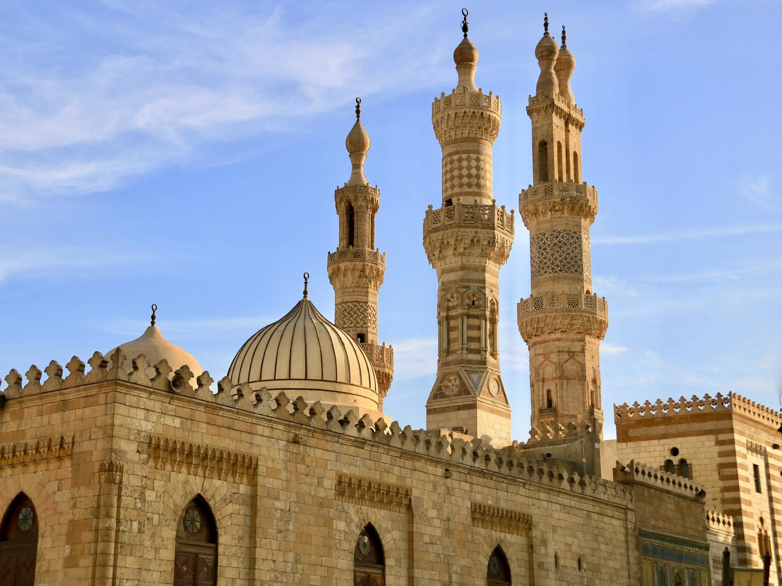 A historic mosque with beige stone walls, domes, and five ornate minarets against a blue sky.