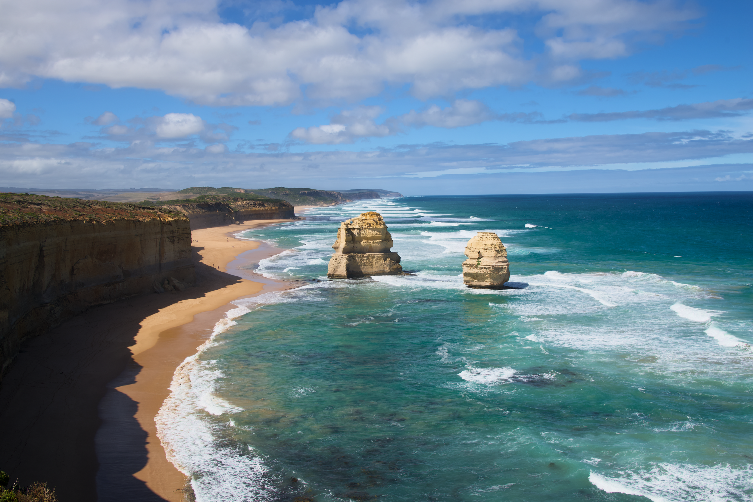 Scenic view of the Twelve Apostles limestone stacks along the Great Ocean Road in Australia, with sandy beaches, green cliffs, and a partly cloudy sky.