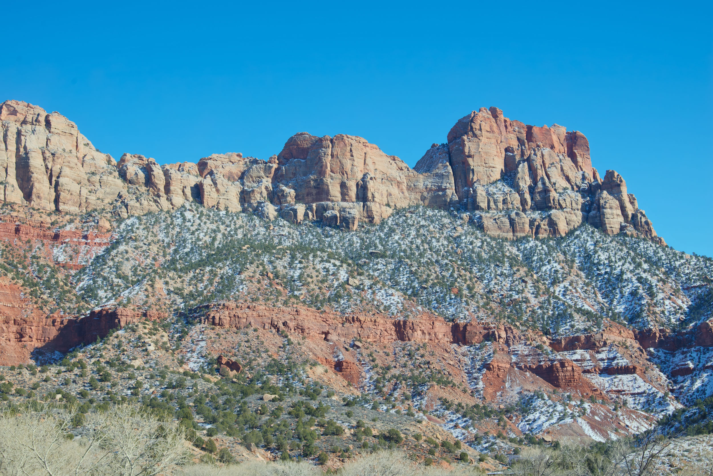 Snow-dusted red rock mountains under a clear blue sky with sparse trees in the foreground.