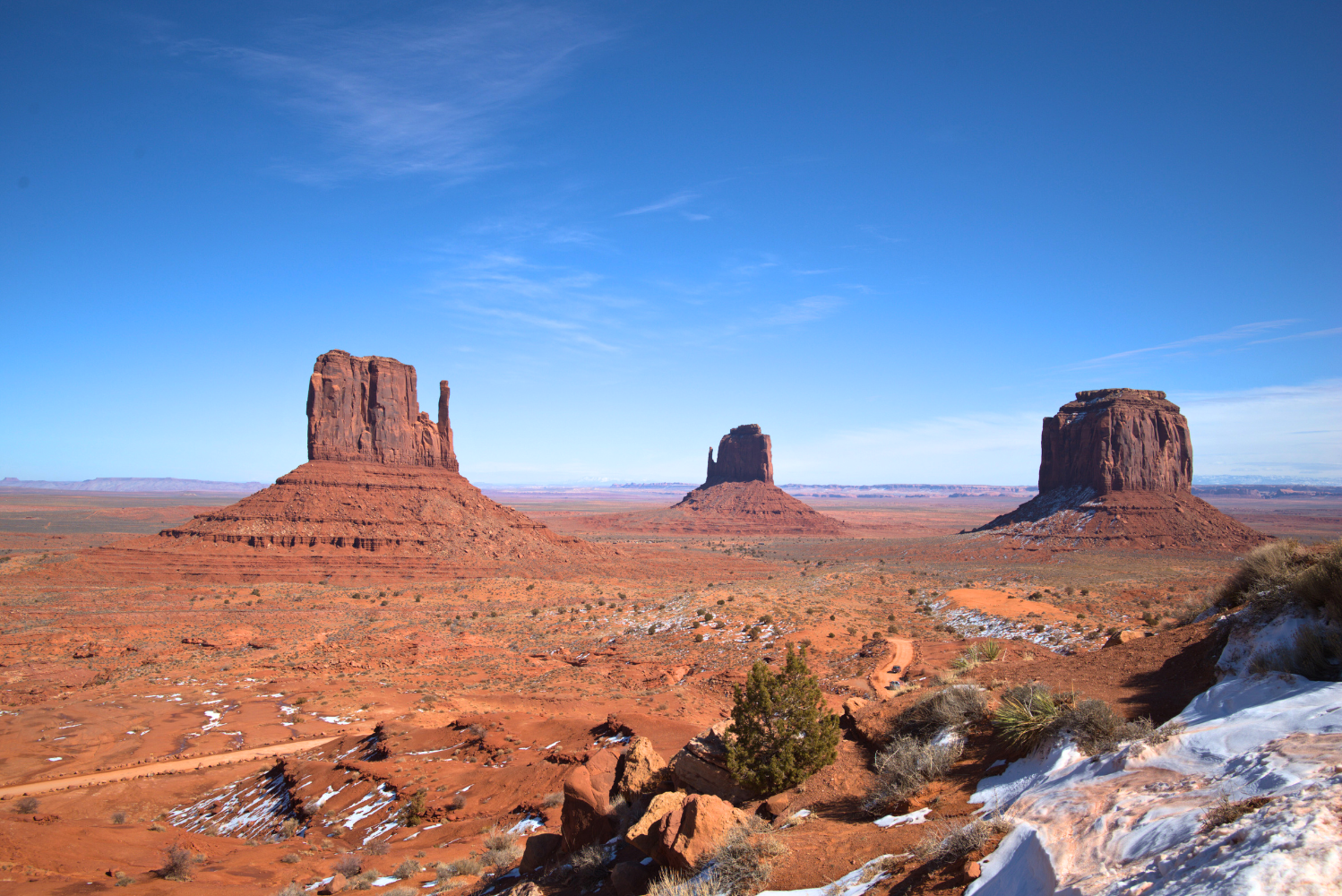 Three large rock formations in Monument Valley, desert landscape with sparse vegetation and patches of snow, under a clear blue sky.