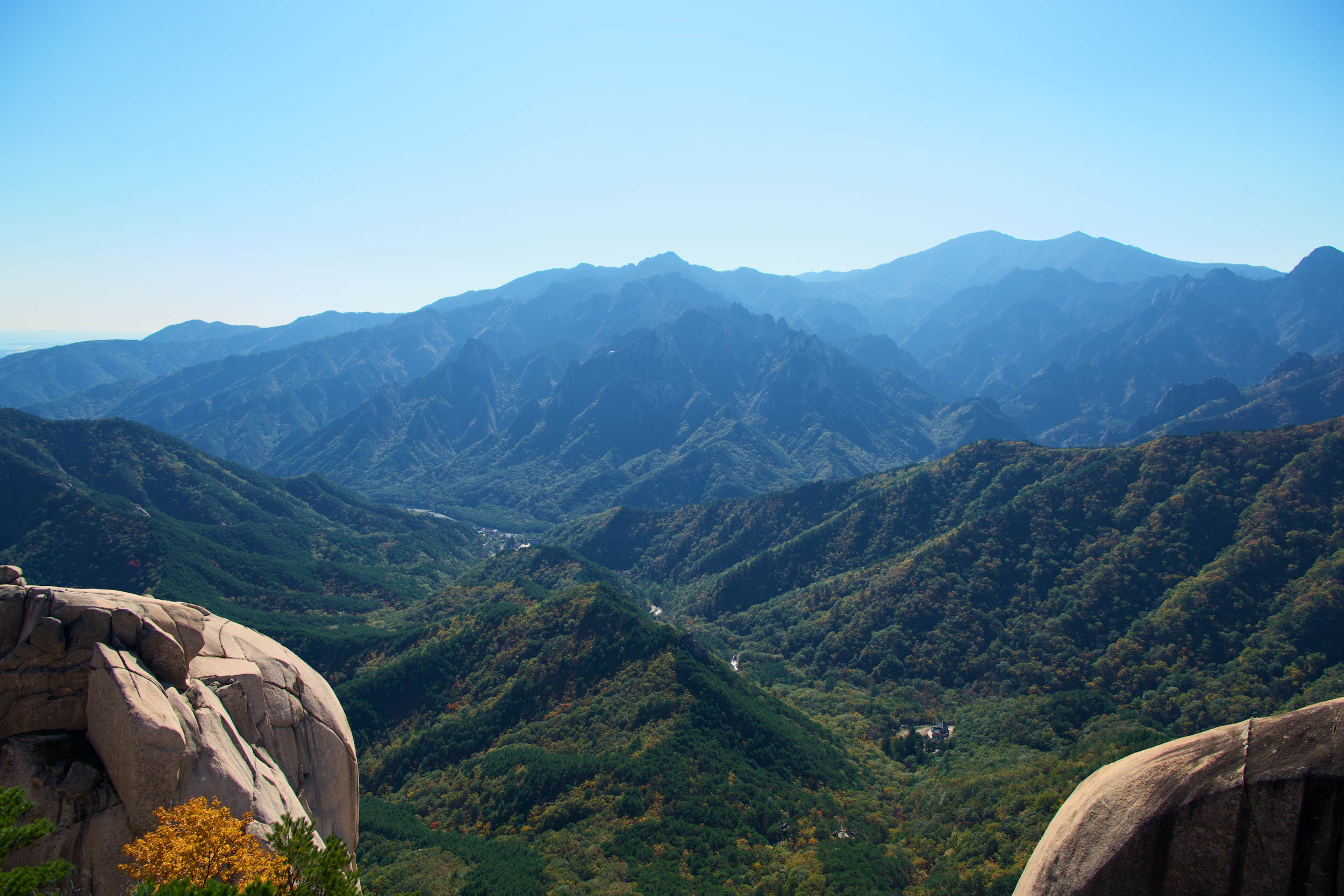 Scenic view of a mountain range with rugged peaks and lush green valleys under a clear blue sky, with large rocks in the foreground.