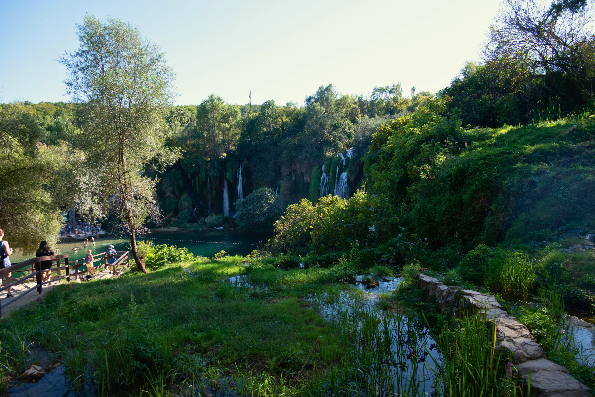 People walking along a path in front of a green landscape with a waterfall in the distance, surrounded by trees and bushes.