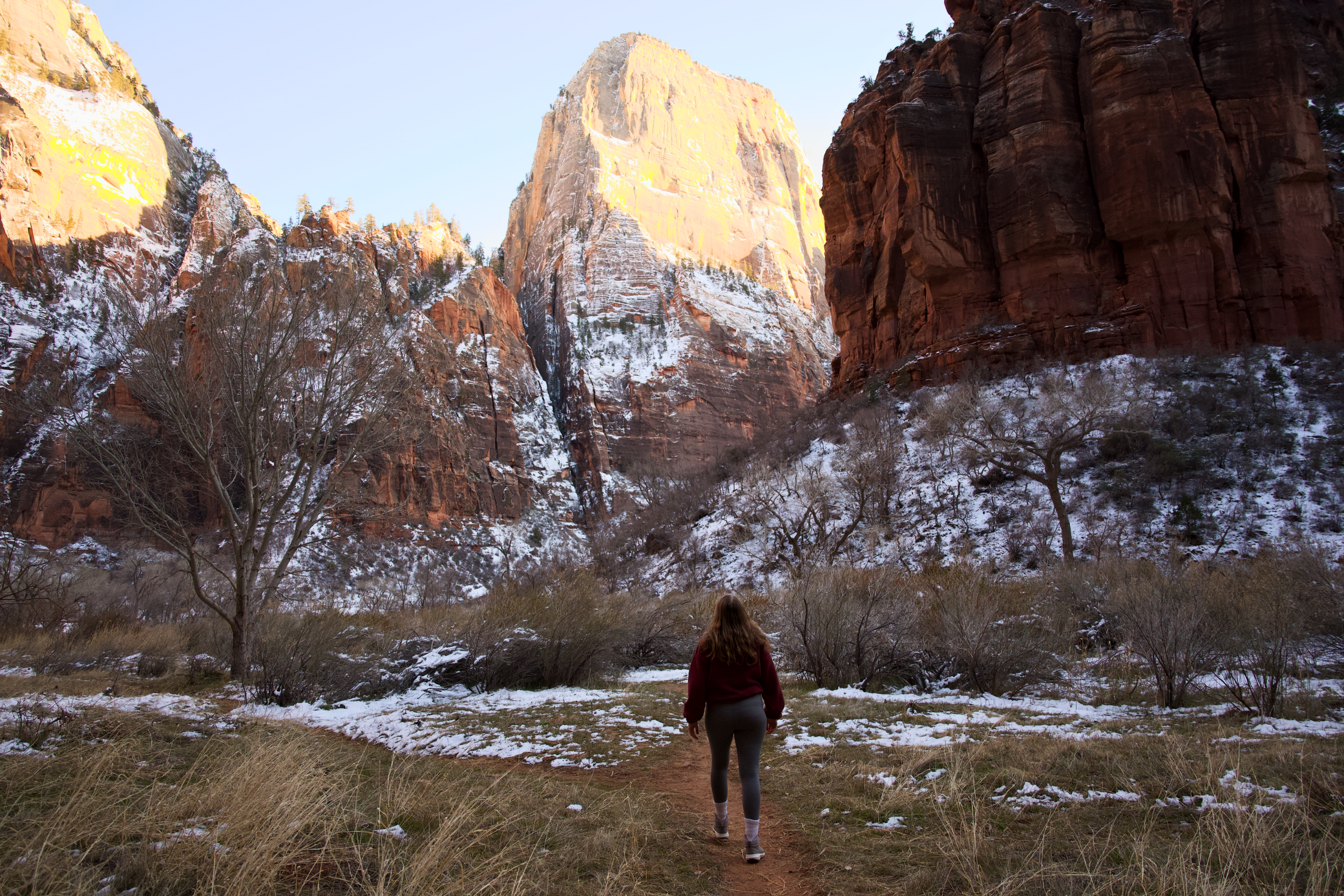 A woman with long hair, wearing a red jacket and gray pants, walking along a dirt trail in a valley surrounded by snow-capped red rock cliffs and leafless trees, during late afternoon or early evening.