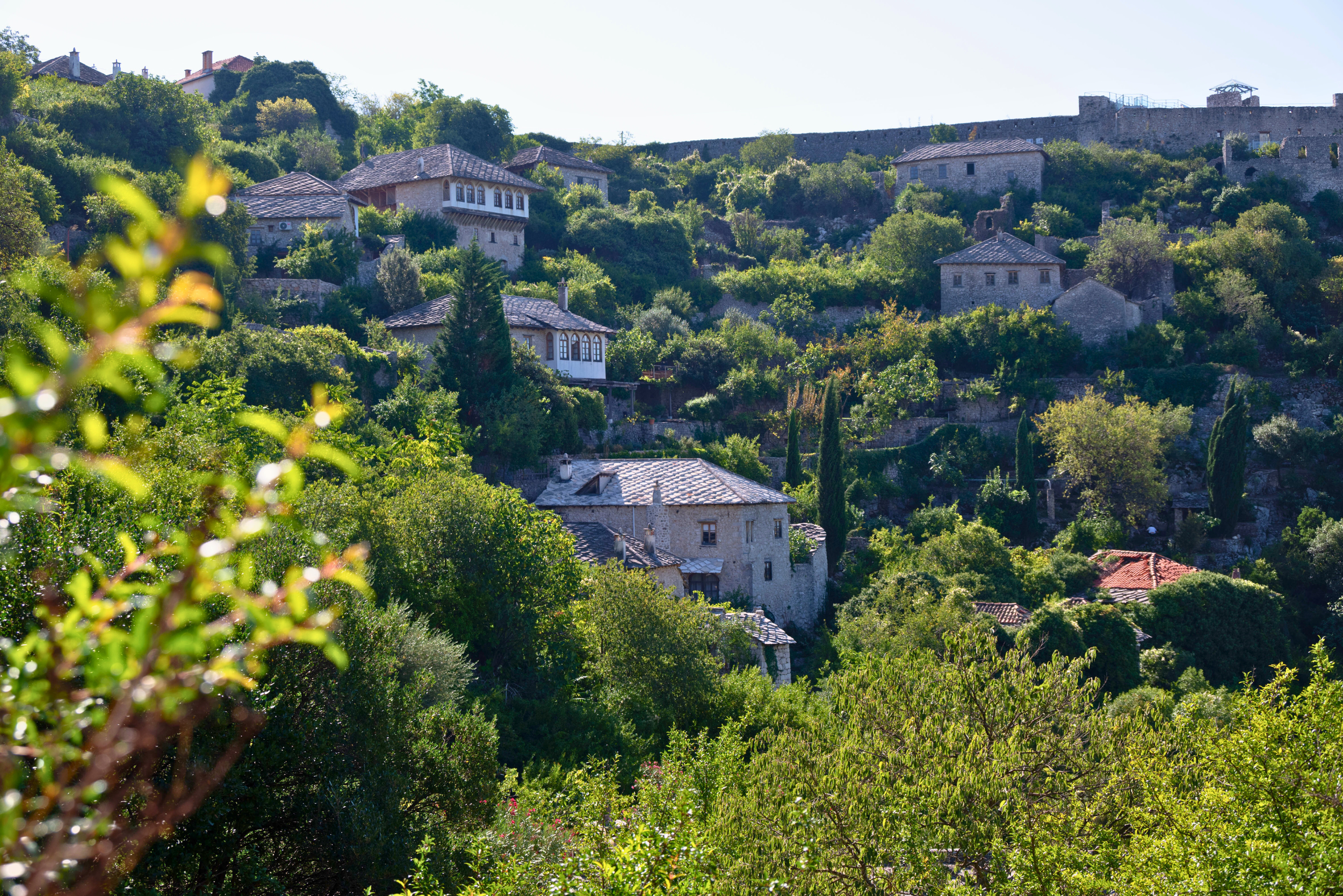 Hilly landscape with numerous stone houses surrounded by green trees and shrubs, part of a historic area with a stone wall at the top.