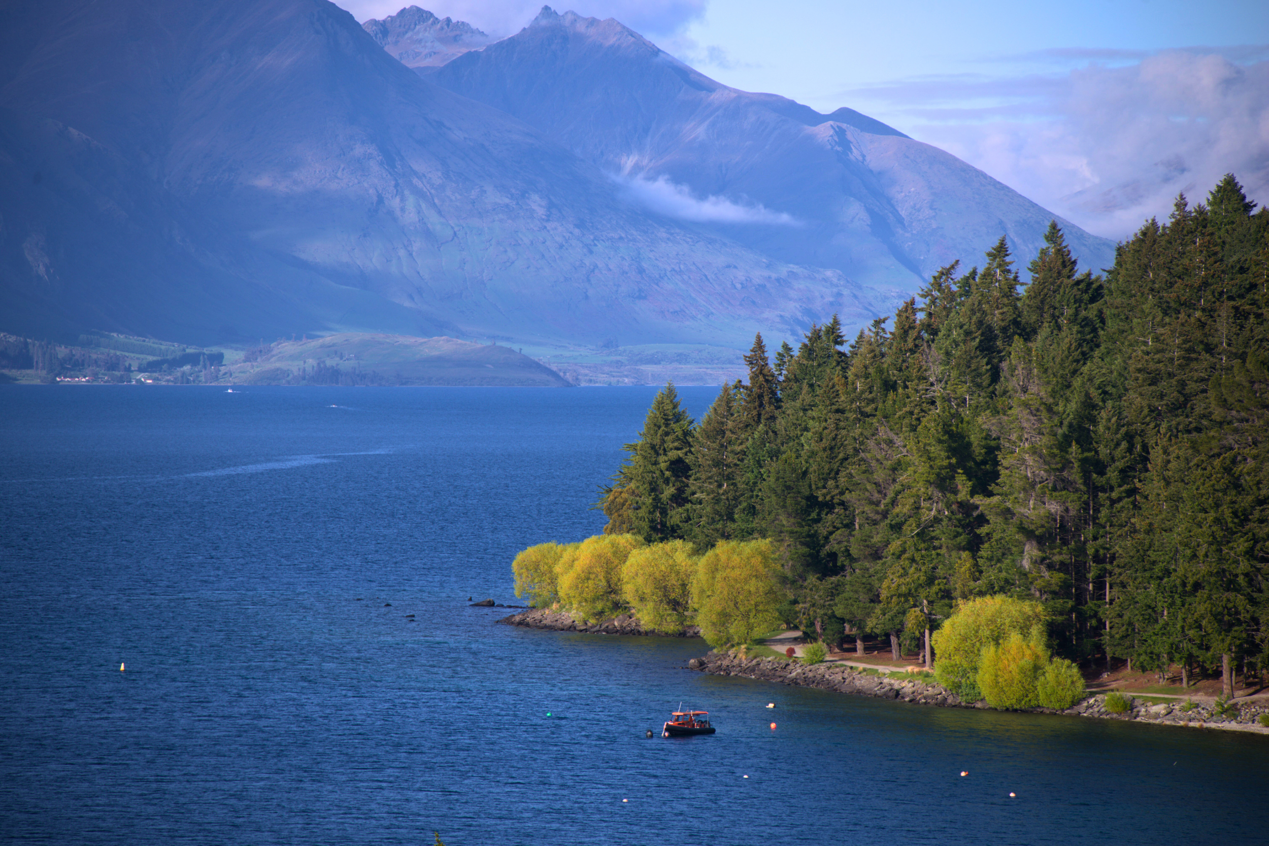 A scenic view of a large blue lake surrounded by green trees on the shoreline and tall mountains in the background with a partly cloudy sky.