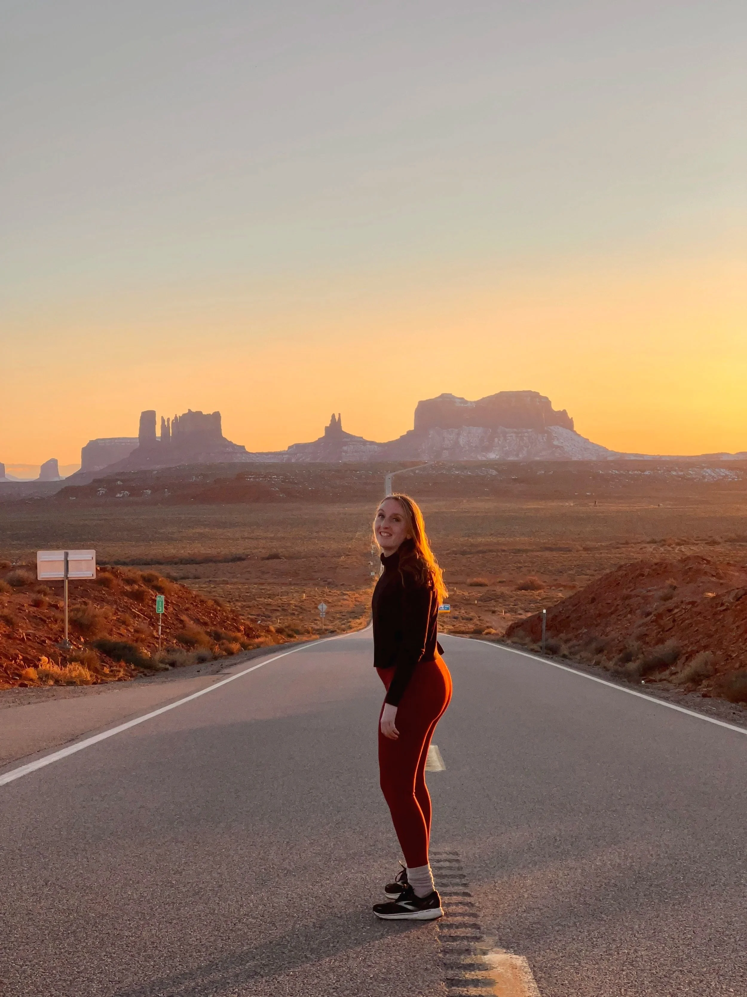 A smiling woman with long hair in athletic clothing standing in the middle of a deserted desert road at sunset, with large rock formations in the background.