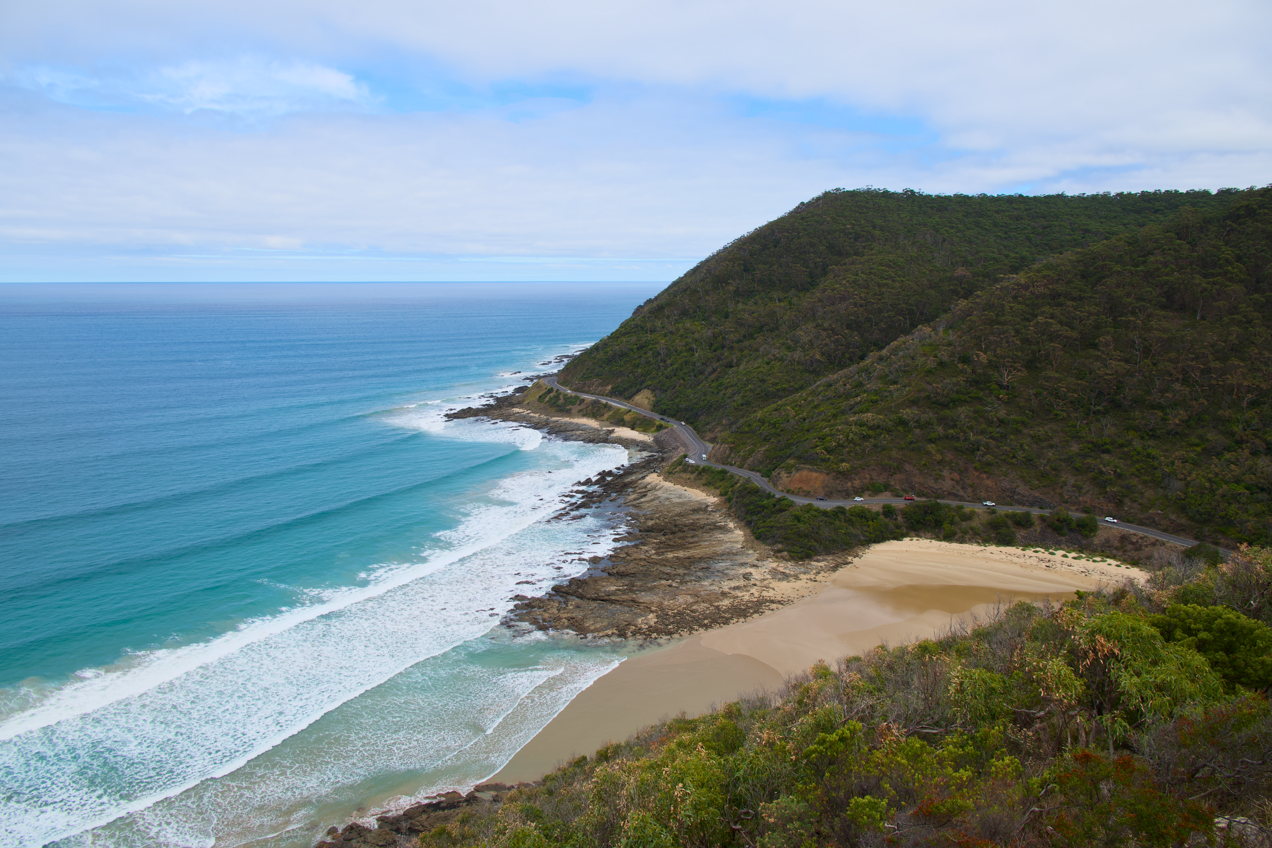 Coastal scene with sandy beach, blue ocean waves, green hillside, and a winding road
