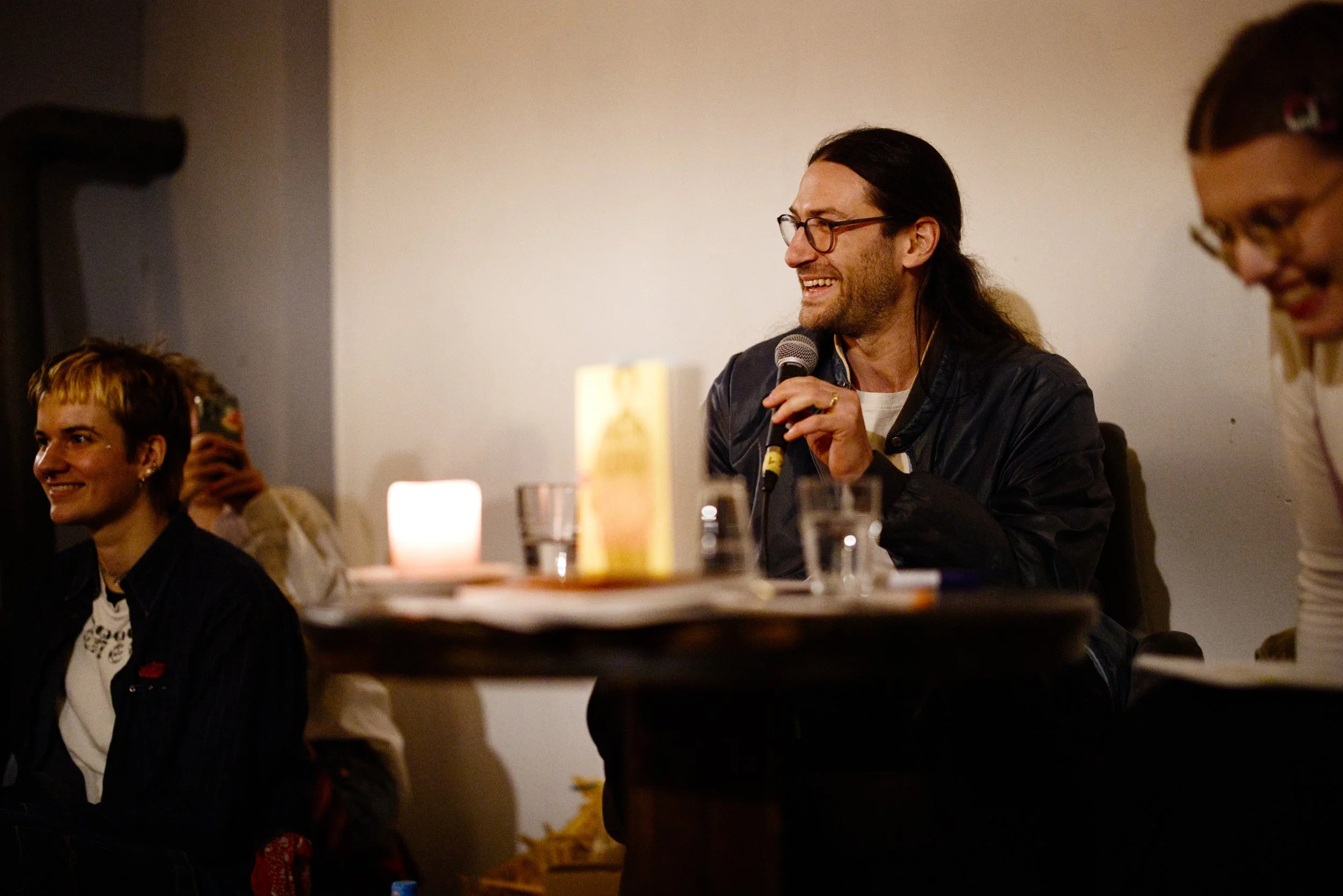 White man with glasses and long brown hair smiling while talking into a handheld microphone. Audre Lorde book in foreground