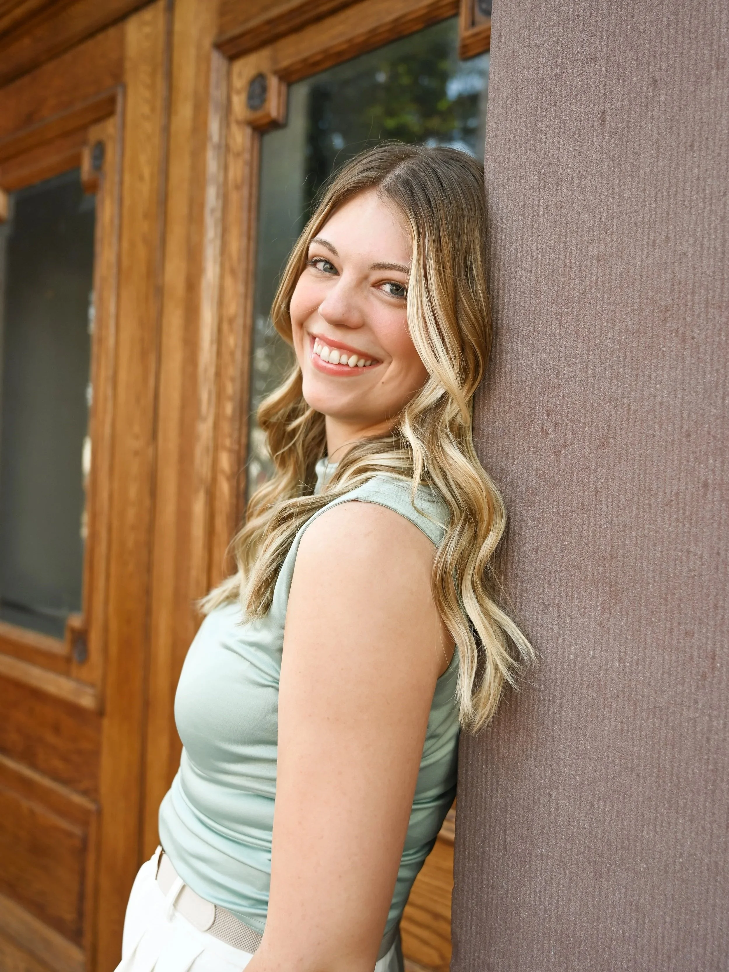 A young woman with long blond hair, smiling, leaning against a wall with wooden window frames behind her.