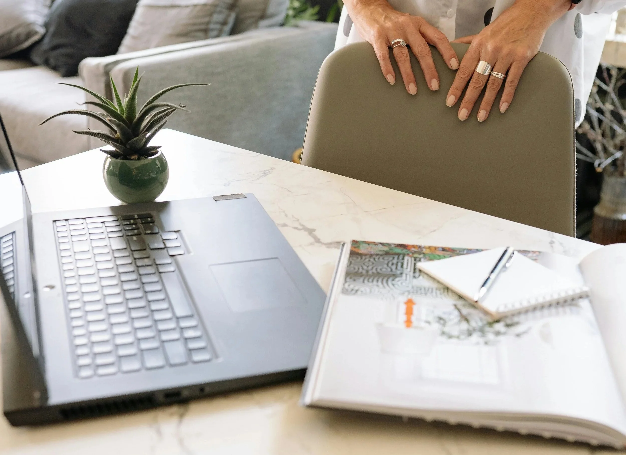 A workspace with a laptop, a potted plant, an open notebook with a pen, and a person's hands resting on the back of a chair. The person is wearing rings on their fingers.