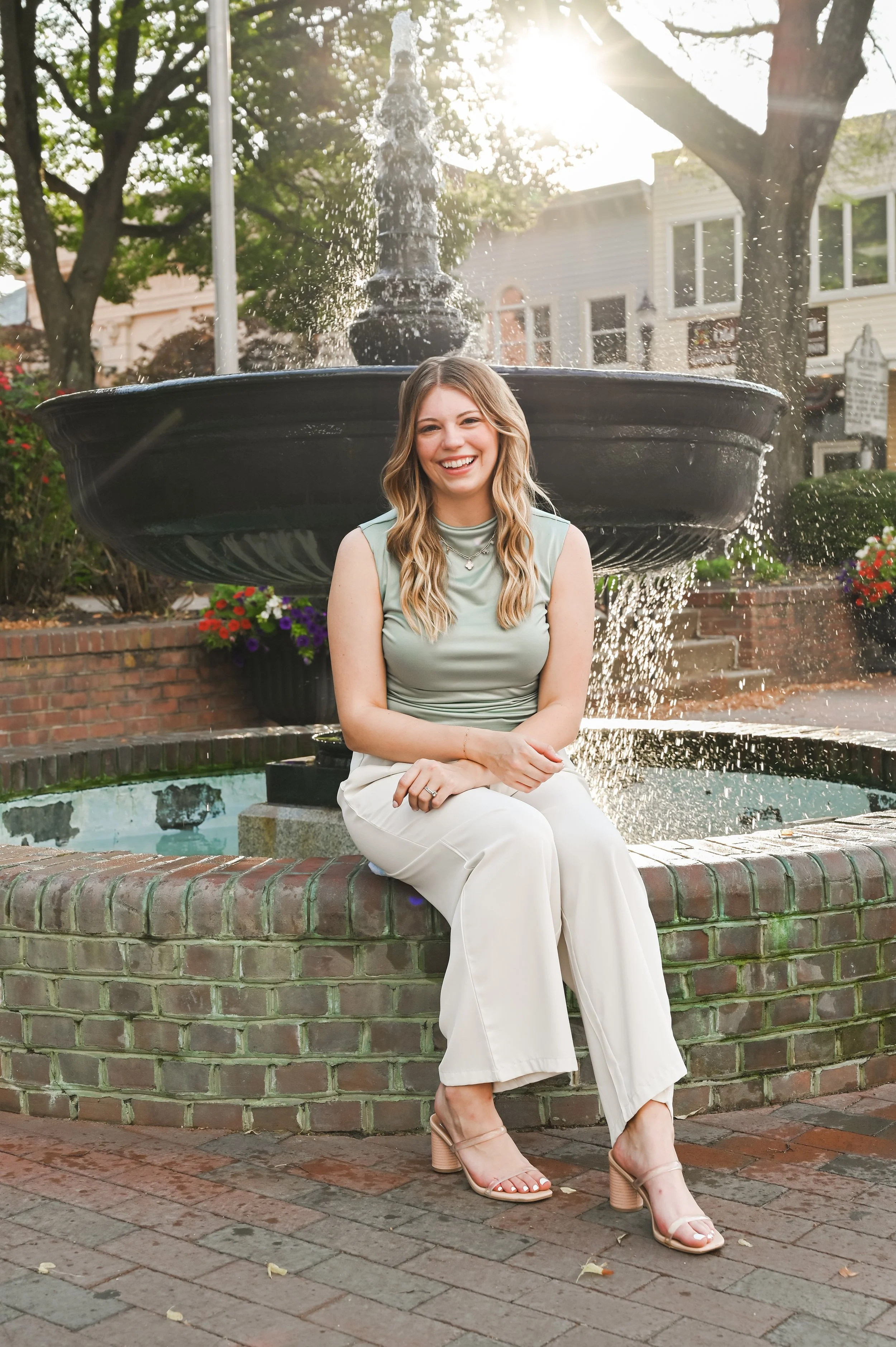 A woman sitting on a brick ledge in front of a fountain, smiling, with a backdrop of trees and houses, during daylight.
