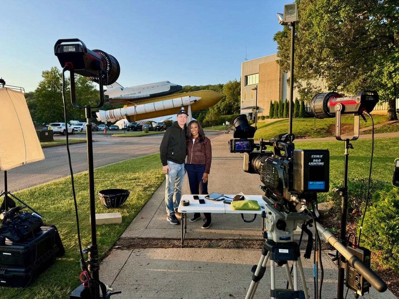 Two people smiling and posing for a photo outdoors at sunset, with spacecraft replicas and a camera setup around them.