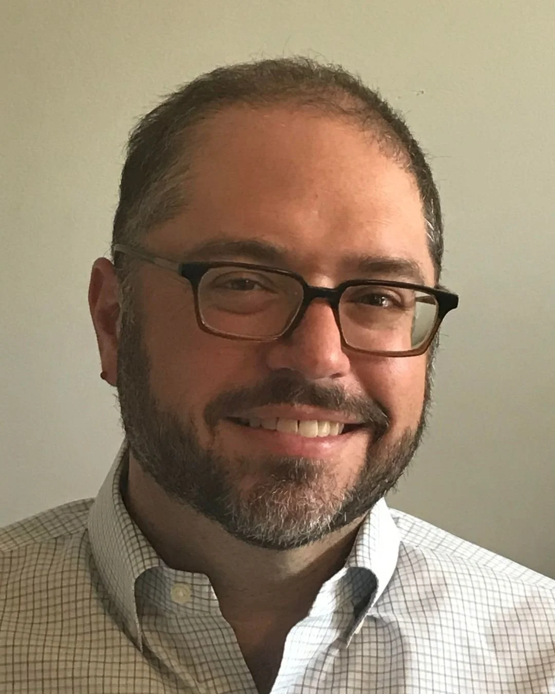 A smiling man with glasses, a beard, and short dark hair, wearing a light-colored checkered shirt, standing against a plain beige wall.