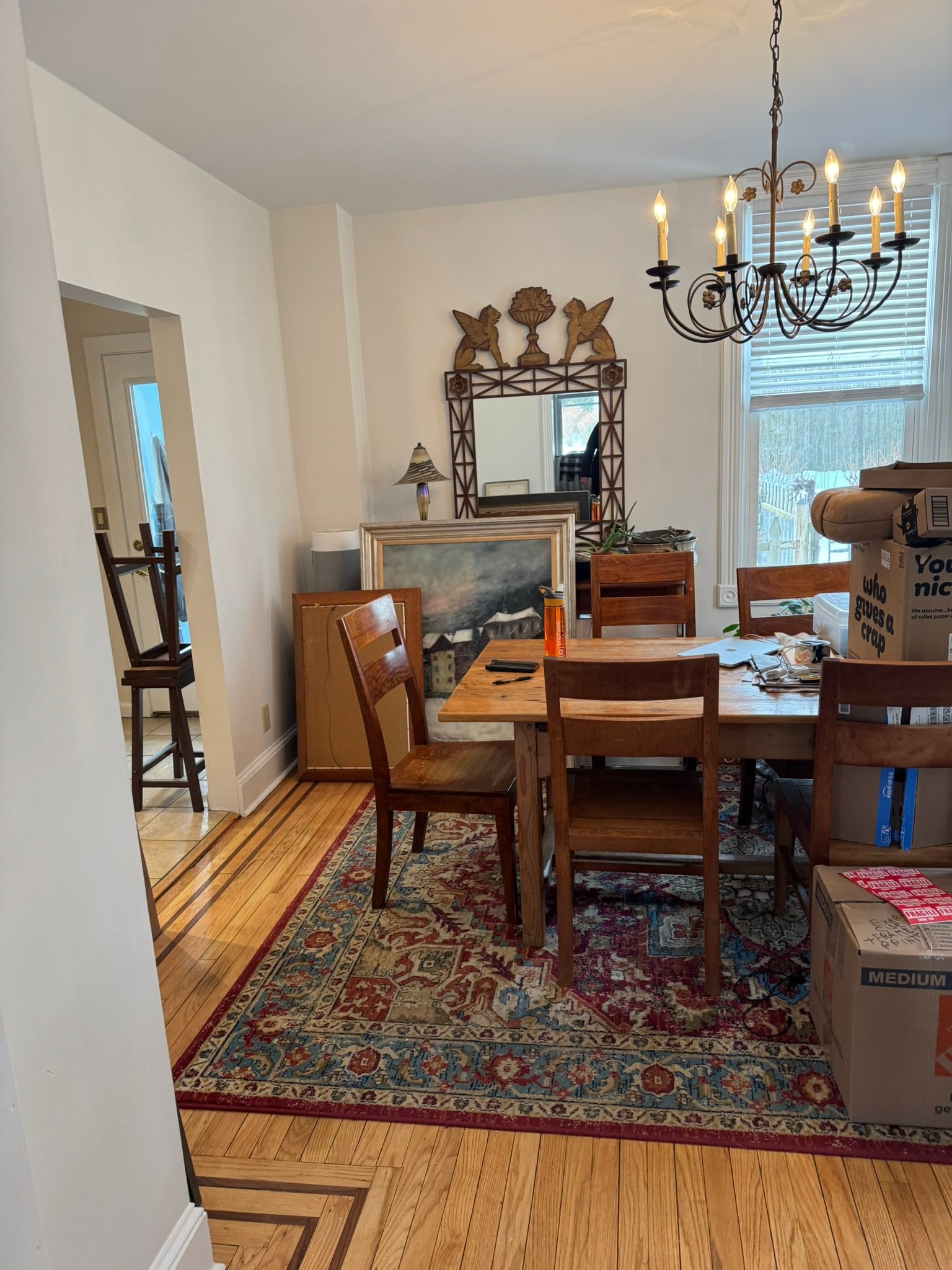 A dining room with a wooden table and chairs on a patterned area rug, a chandelier hanging from the ceiling, and a wall with window blinds, a mirror, and decorative shelves with cherub and bird carvings. Items on the table include a black object, pap