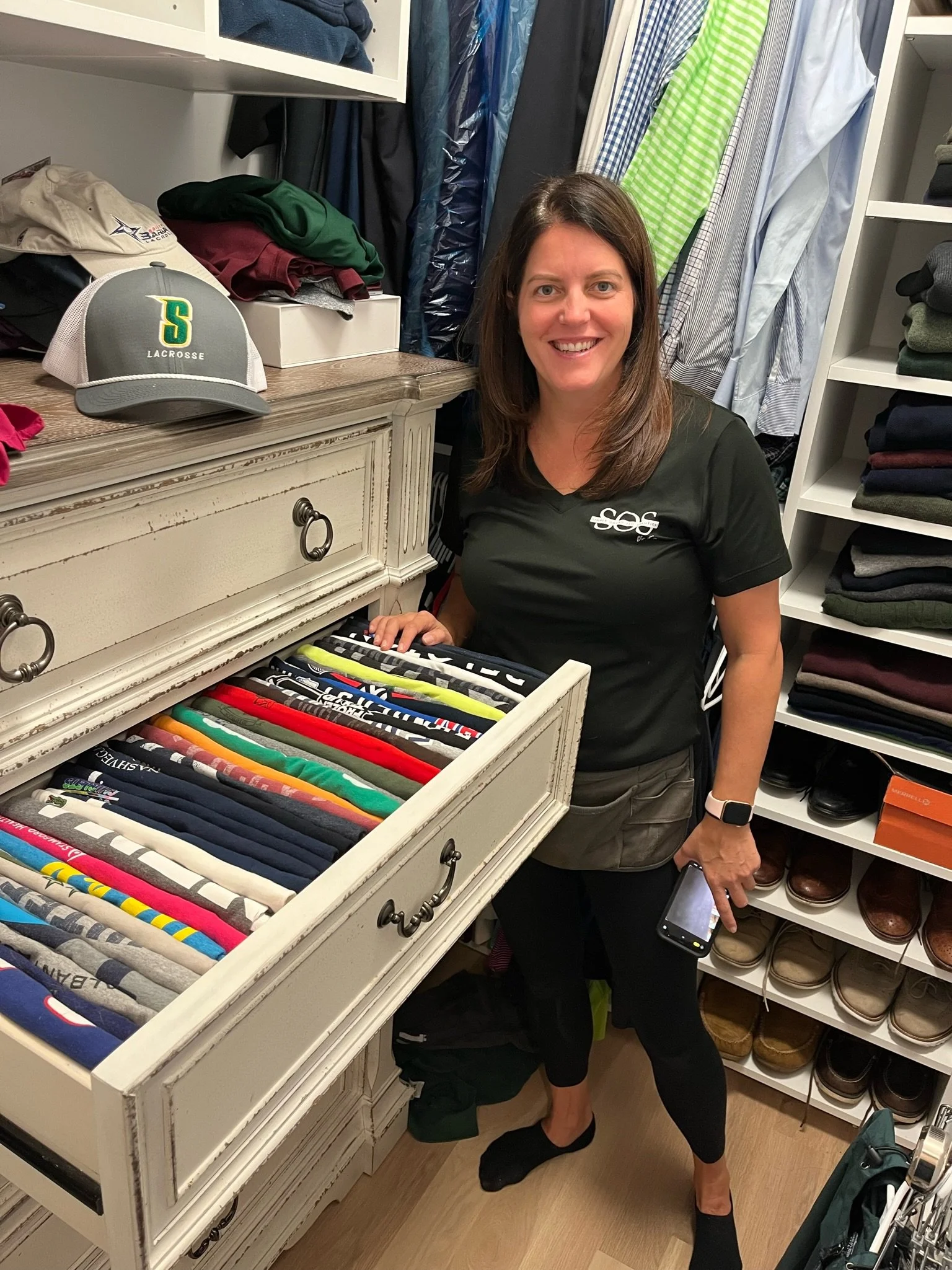 A woman with shoulder-length brown hair, wearing a black t-shirt, and holding a smartphone, standing in a closet or dressing room. She is smiling and leaning slightly on a white dresser drawer filled with neatly folded T-shirts, with various clothing items and shoes on shelves around her.