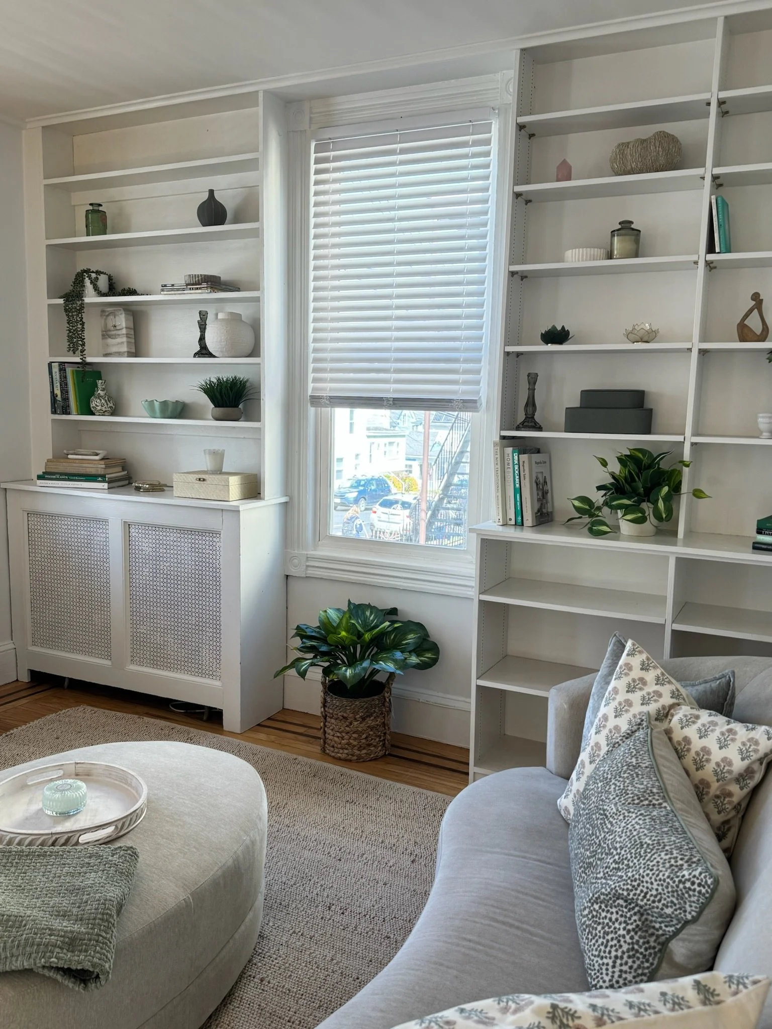 Living room with white bookshelf, window with white blinds, green plants, decorative pillows on a curved sofa, and a white ottoman with a tray and candles.